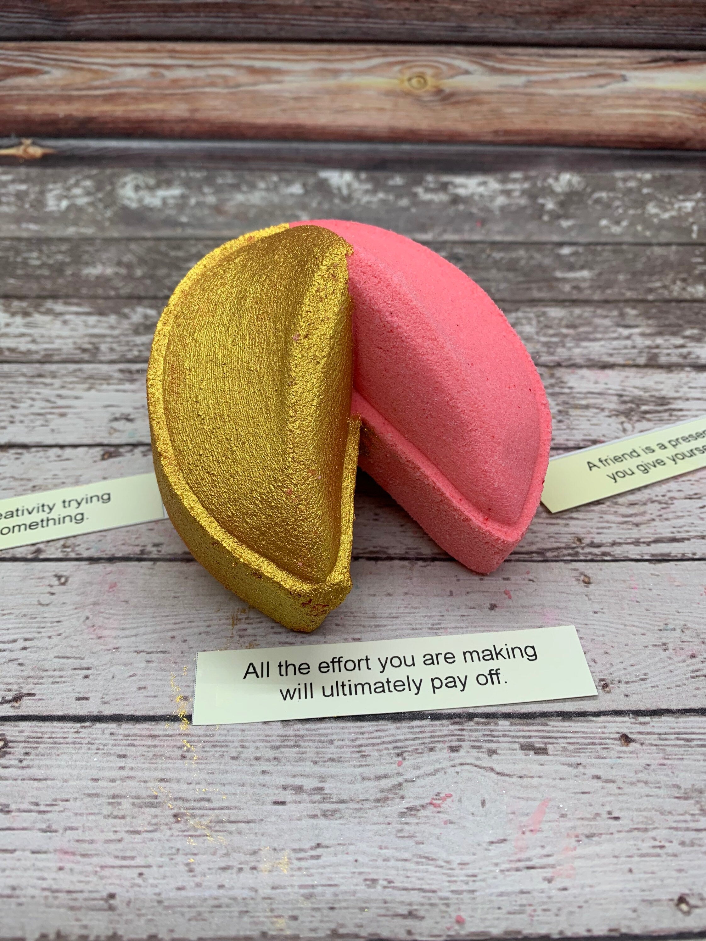 One pink fortune cookie shaped bath bomb. Half is hand-painted gold shimmer. A fortune saying "All the effort you are making will ultimately pay off" is displayed in front.