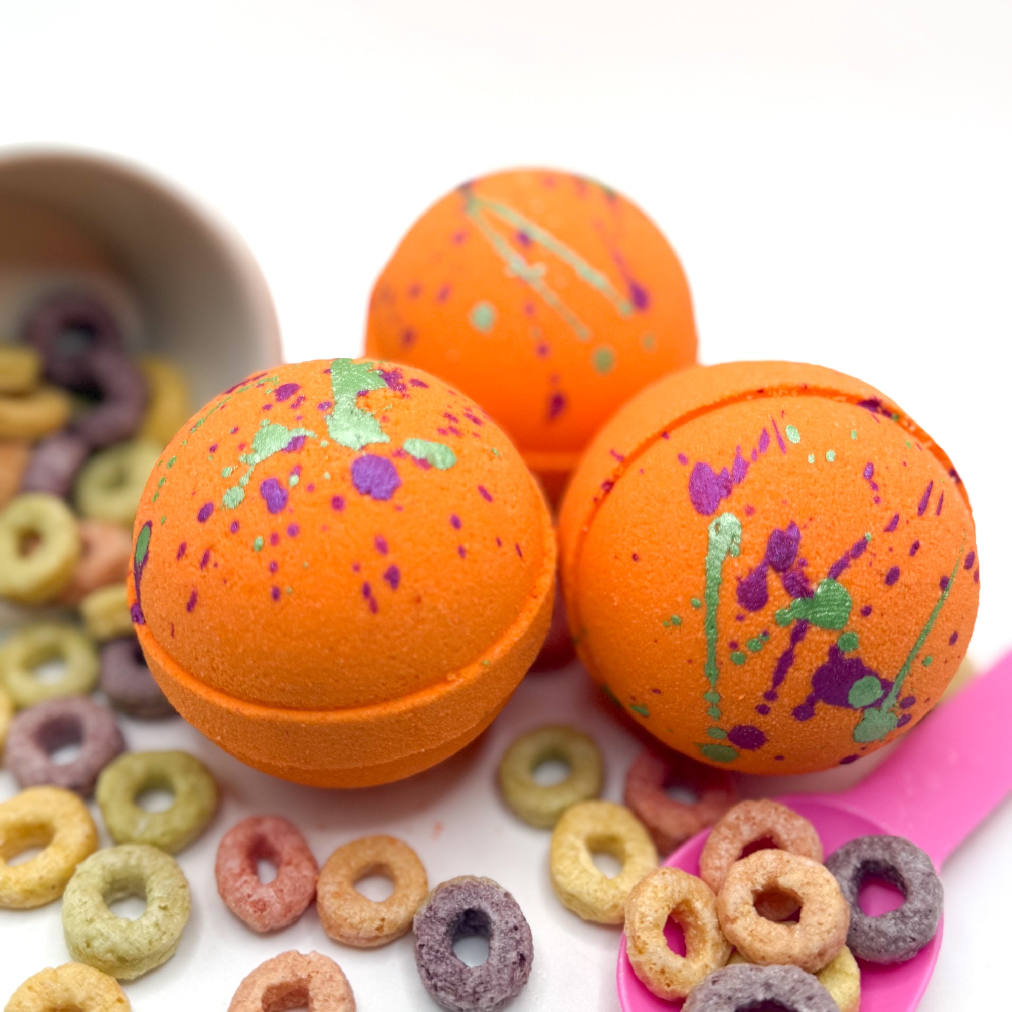 Three orange fruity cereal bath bomb with hand painted green and pink specks on top. Pictured on a white background with cereal loops scattered about