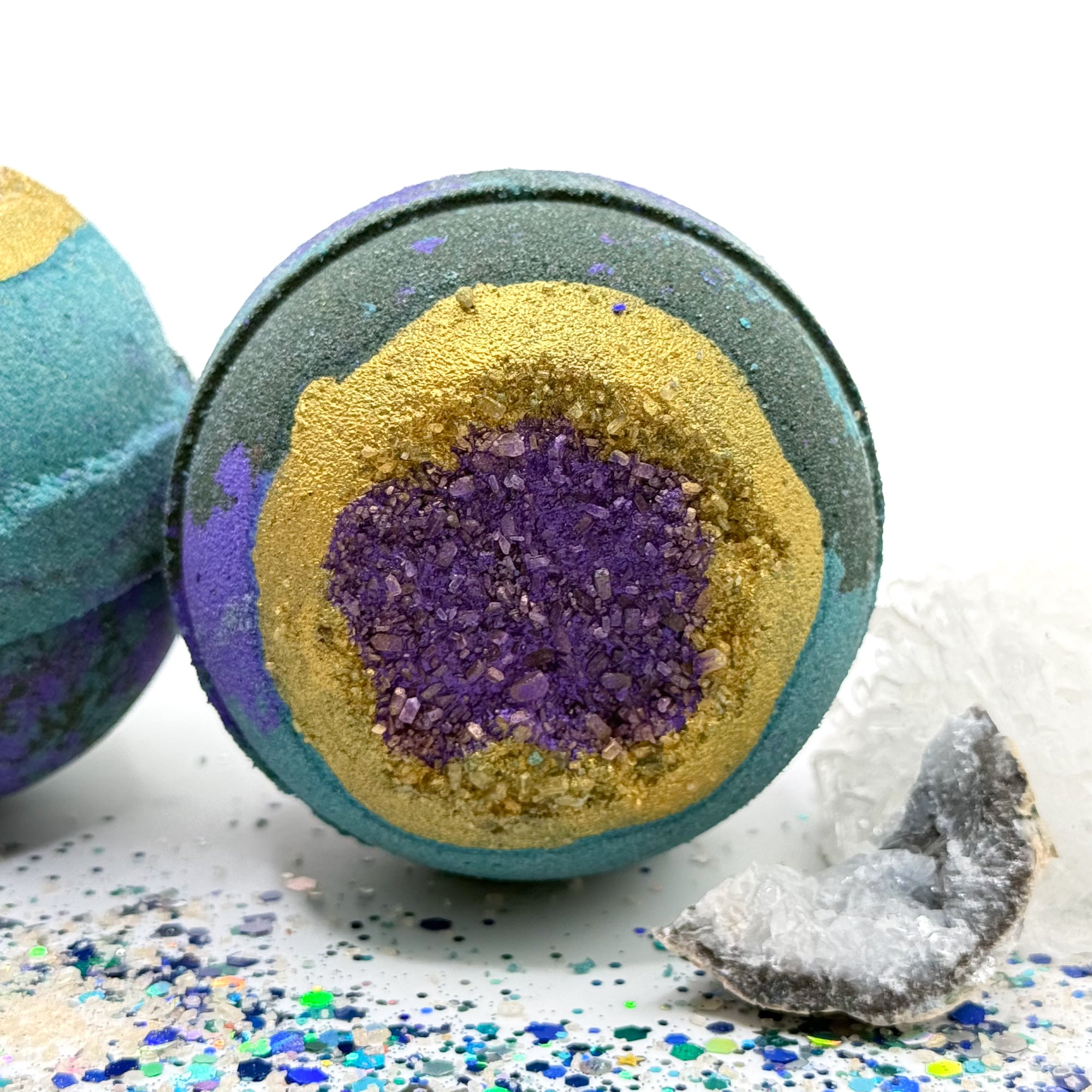 Two green, blue and purple bath bombs one facing forward and the other one facing upright with purple and gold centers resembling a geode on a glittery surface with crystals.