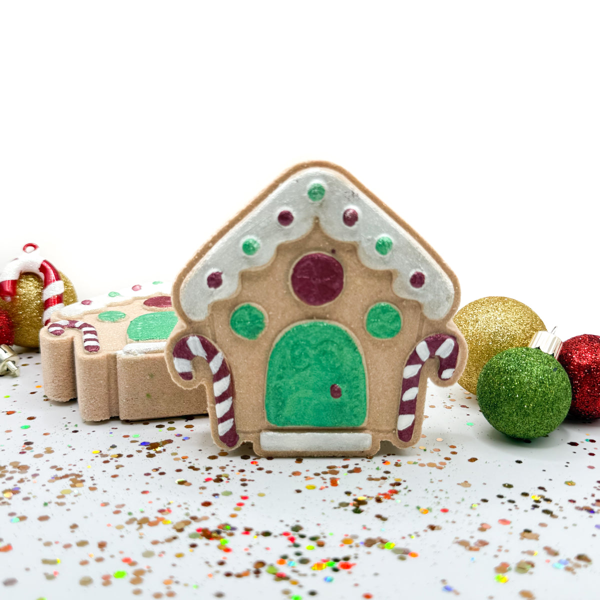 Front and side view of hand painted gingerbread house bath bombs with Christmas ornaments in the background.