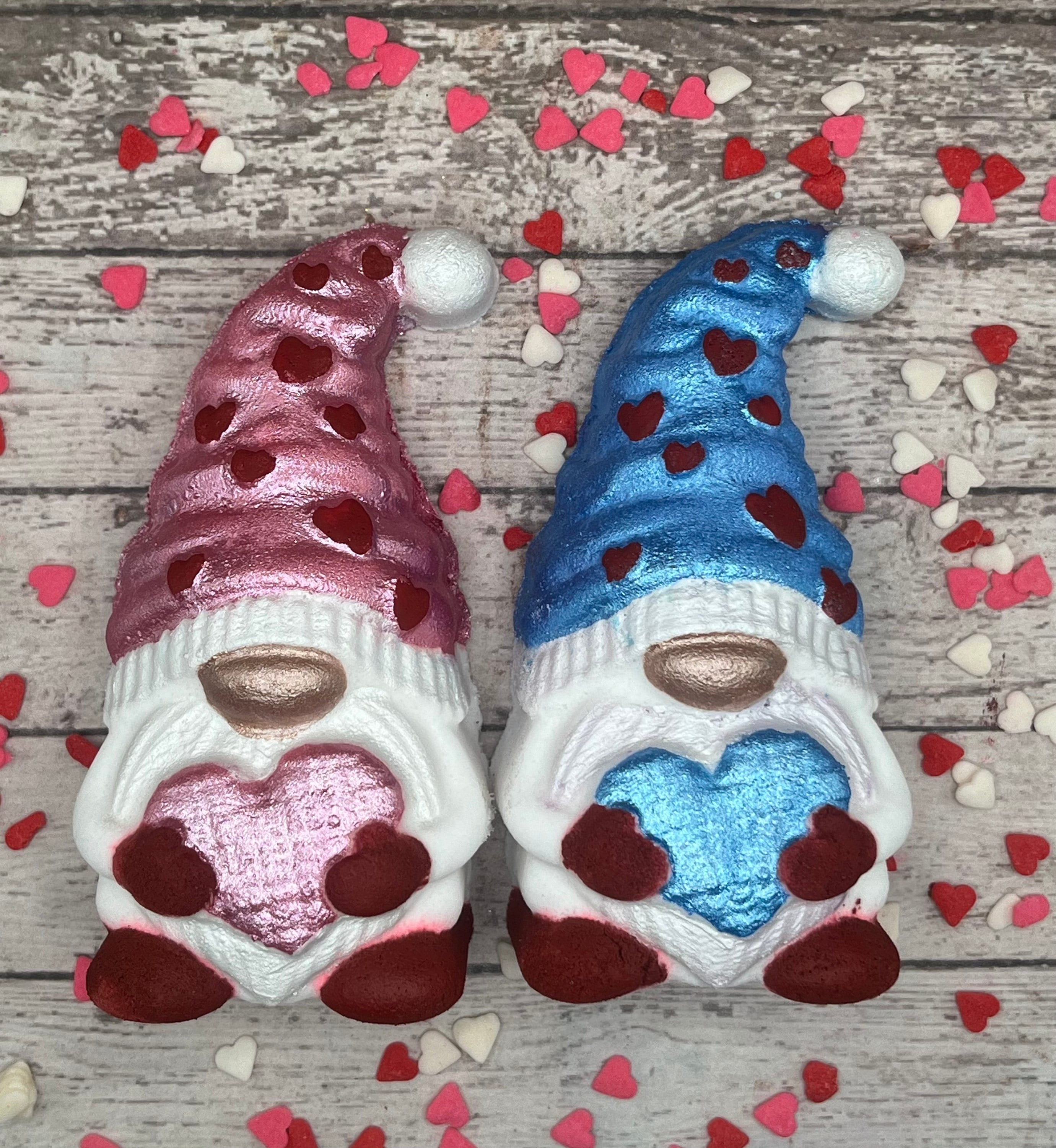 2 gnome shaped bath bombs holding hearts. One is holding a pink heart with red heart accents the other is holding a blue heart with blue hat and red heart accents.