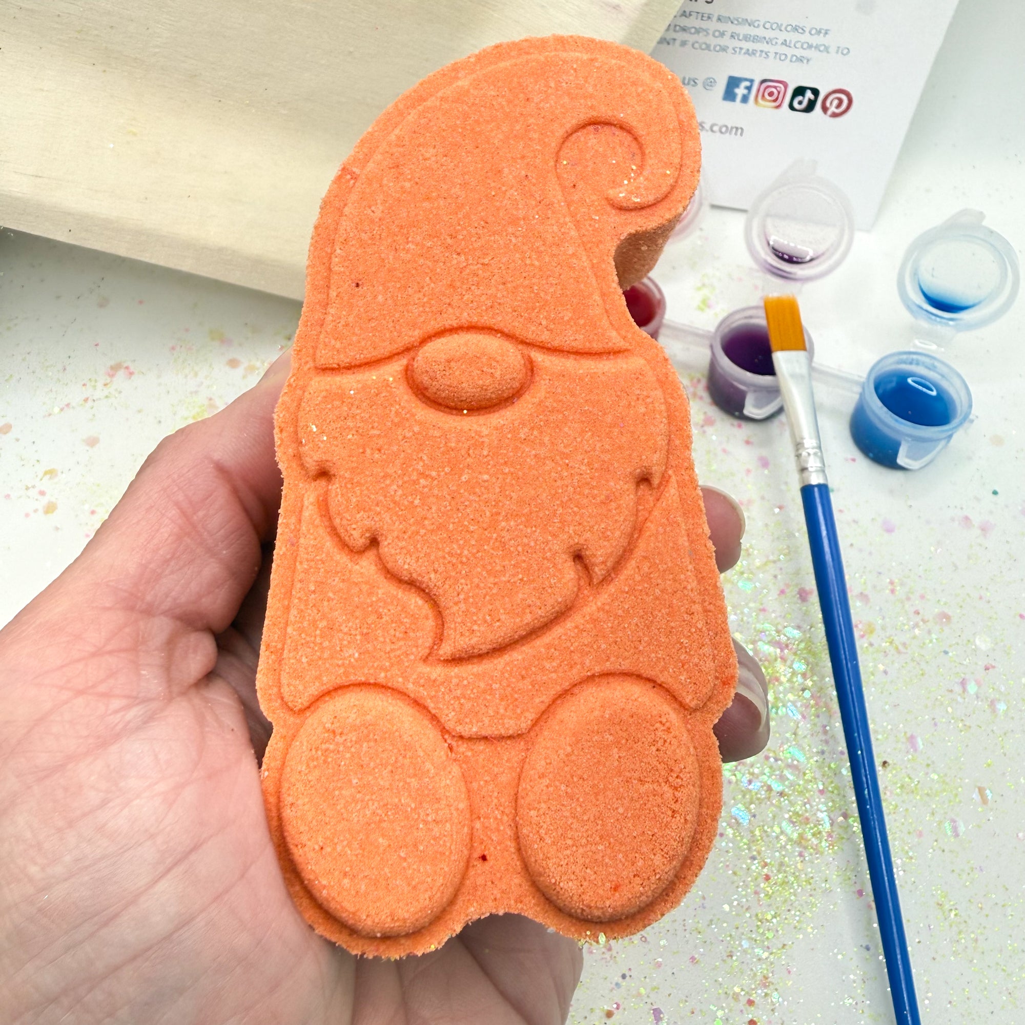 Orange paint your own gnome bath bomb held by a hand with paint supplies in the background