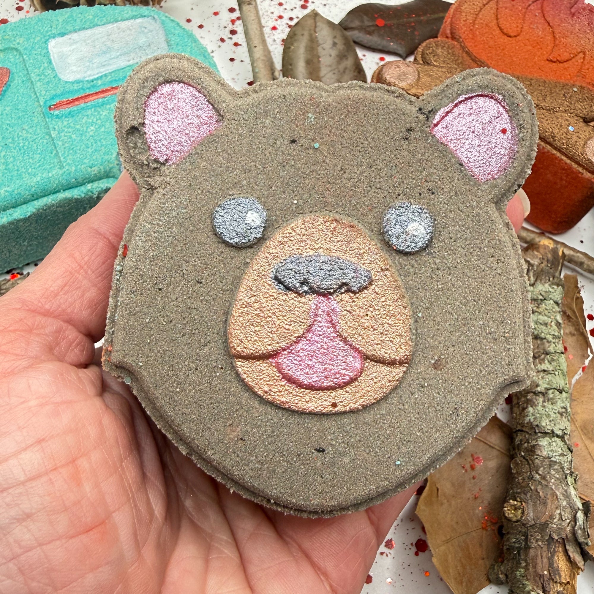 Hand holding a hand painted brown bear shaped bath bomb with glittery eyes and pink tongue against a background of other soaps.