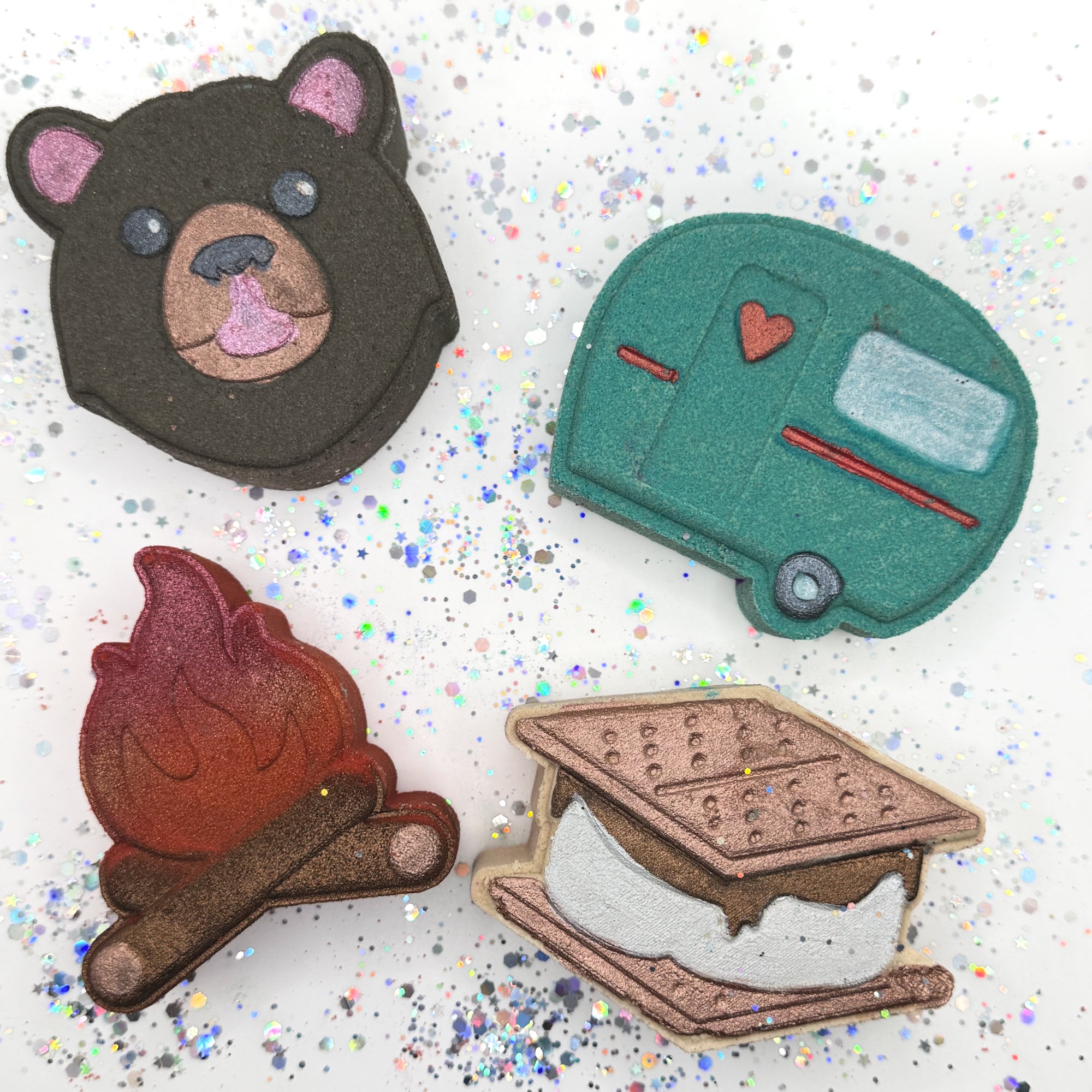 Four handmade bath bombs on a glittery background: bear, camper, campfire, and s'mores.