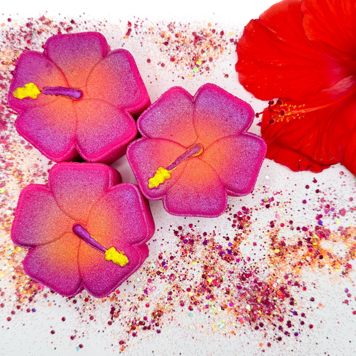 Three hibiscus shaped bath bombs with an airbrushed pink-to-orange gradient and yellow stamen details, arranged on a white surface with scattered glitter and a red hibiscus flower prop.