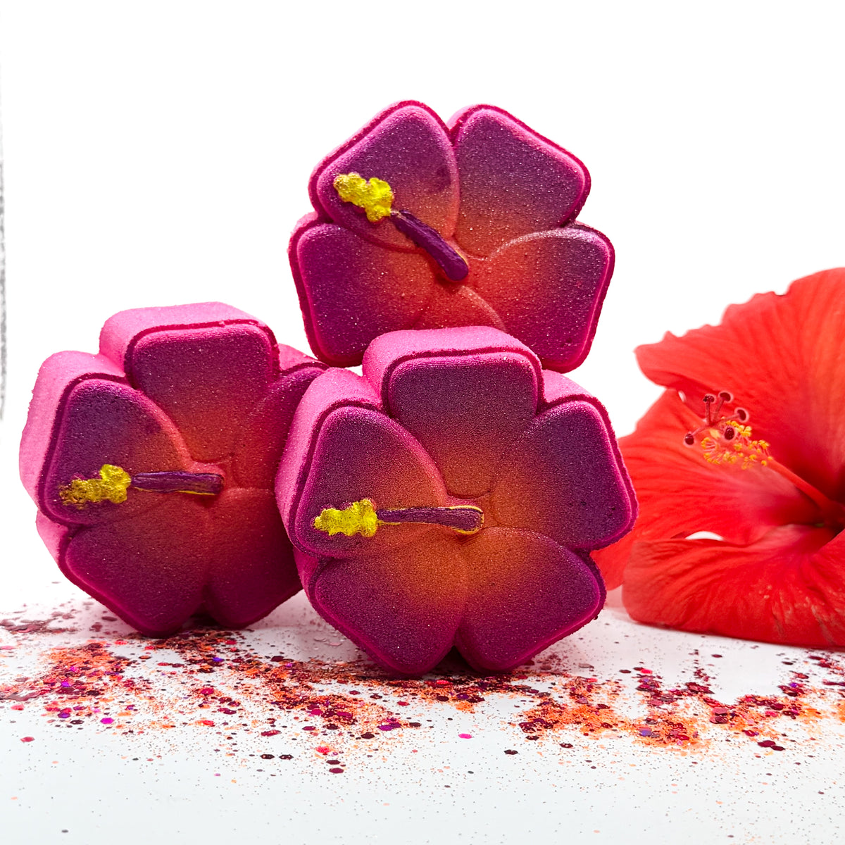 Three hibiscus shaped bath bombs with a pink-to-orange gradient and yellow stamen details, arranged on a white surface with scattered glitter and a red hibiscus flower prop.