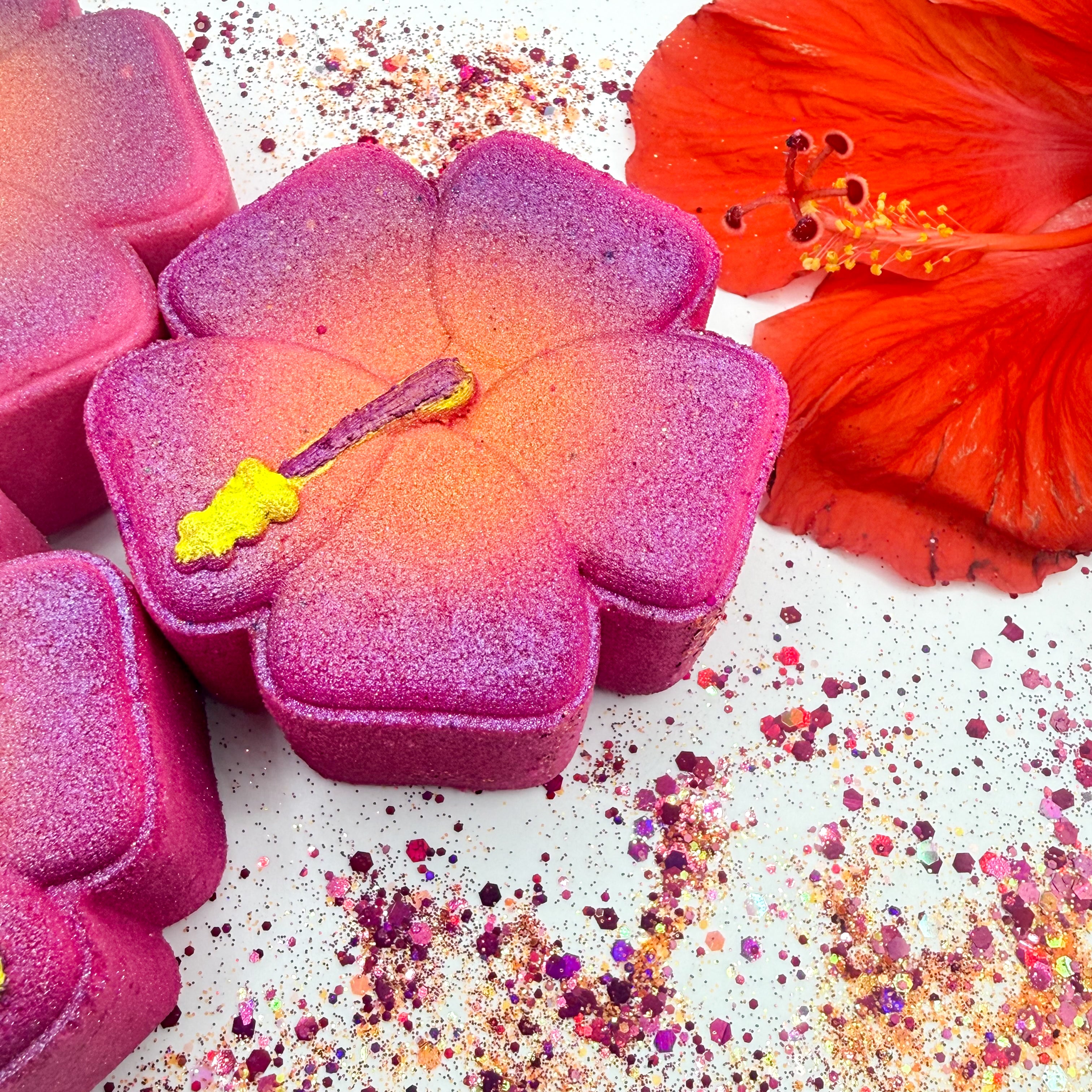 A close up flat lay of hibiscus shaped bath bombs in a glittery magenta-to-pink gradient, with a bright orange hibiscus flower and scattered multicolor glitter on a white background.