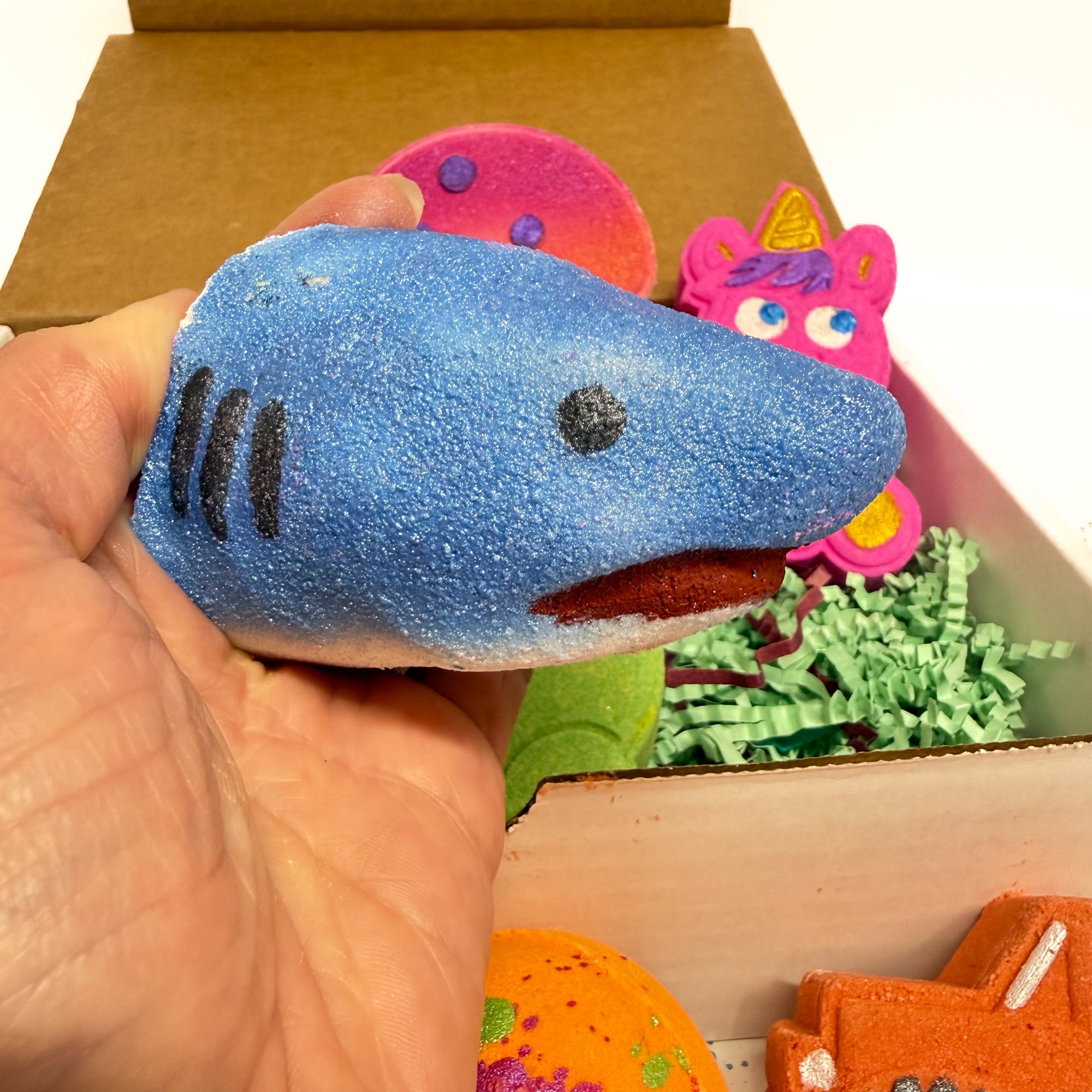 Blue shark bath bomb being held in a hand with other colorful bath bombs in the background.