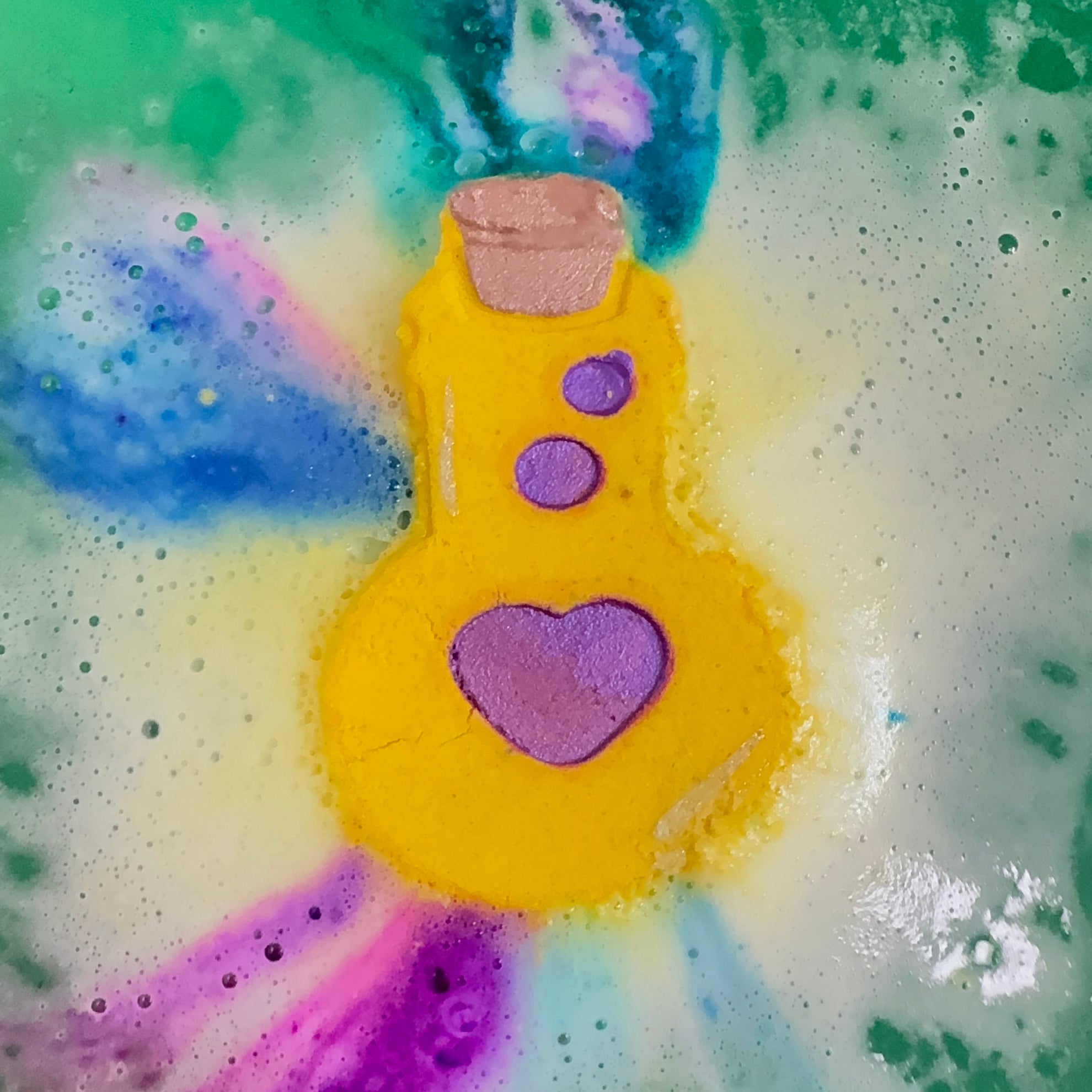 Yellow love potion bath bomb with a purple heart design and colorful colors coming out in a bathtub filled with bubbles and colorful water.