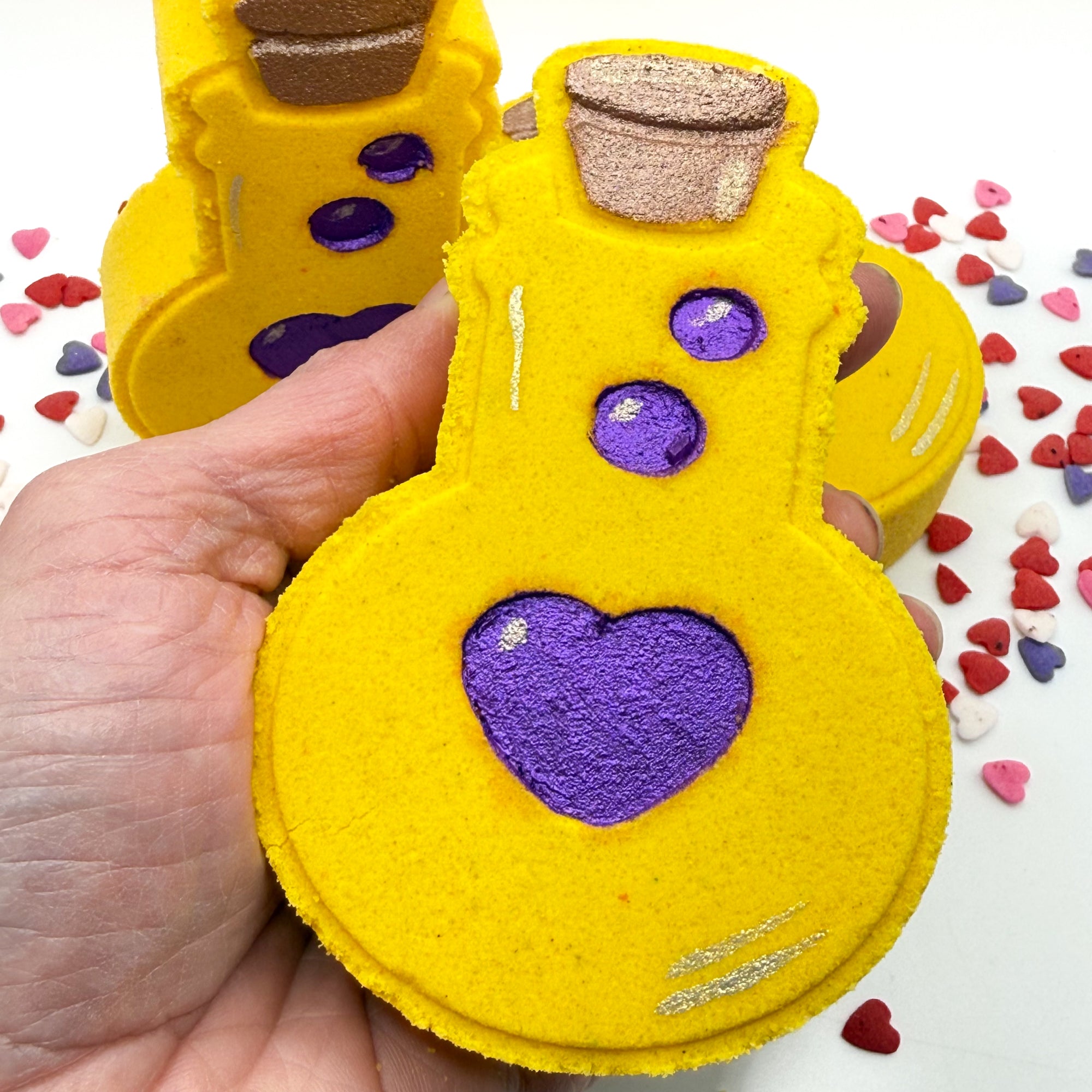 Hand made yellow love potion bath bomb with purple heart held in a hand, surrounded by colorful heart-shaped candies.