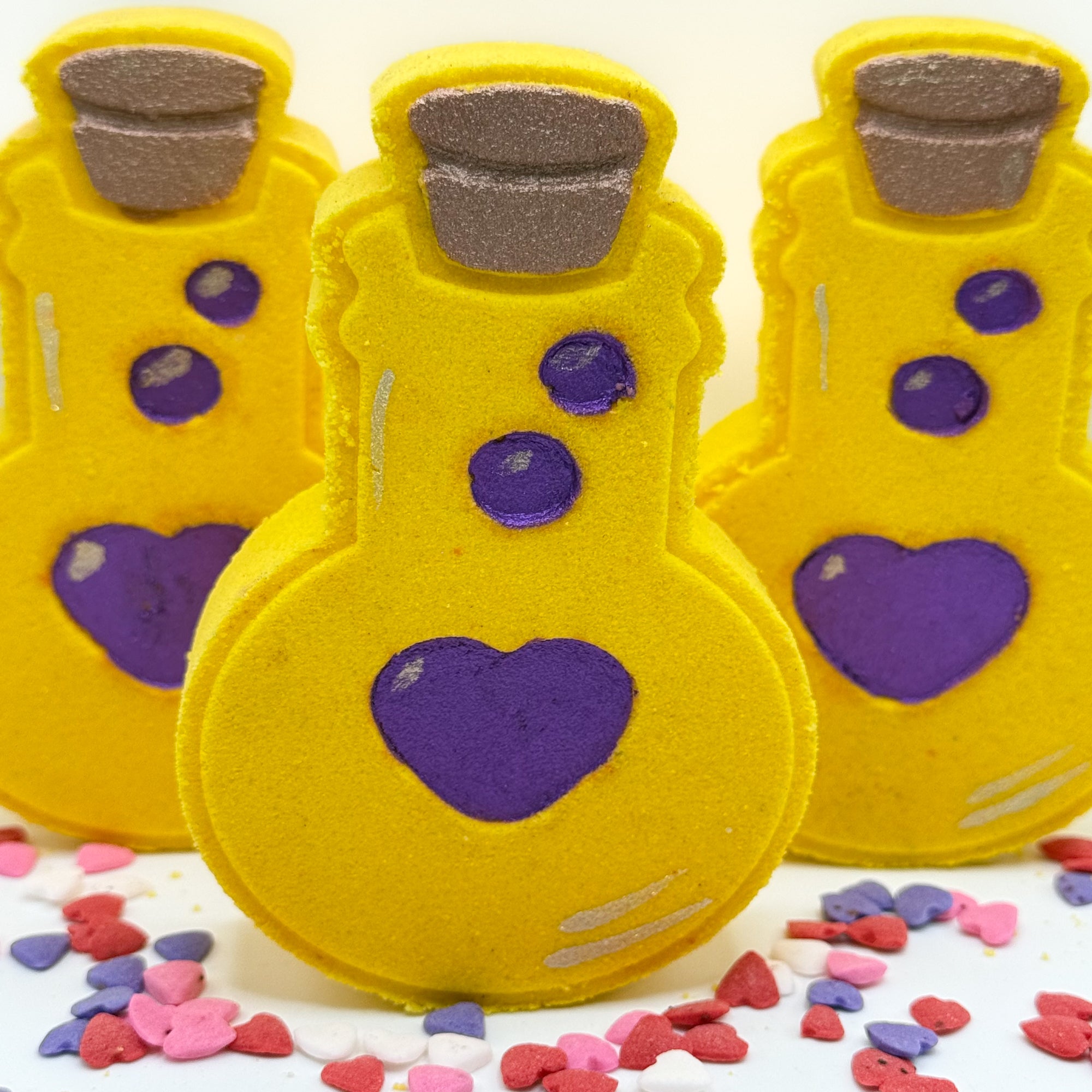 Three yellow love potion bottles bath bombs with purple hearts on a white background. Hand made by Rusted Acre Soap Co.