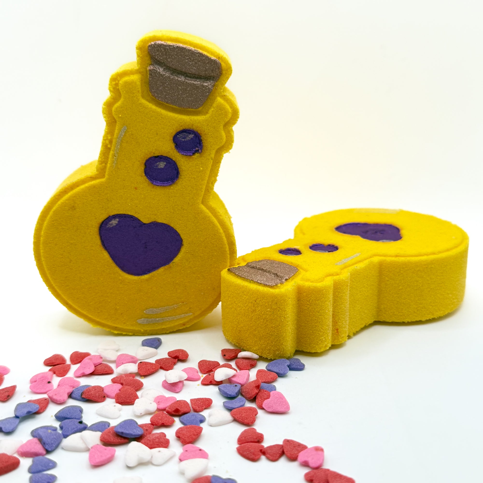 Two yellow love potion bottles bath bombs with purple hearts on a white background. Hand made by Rusted Acre Soap Co.