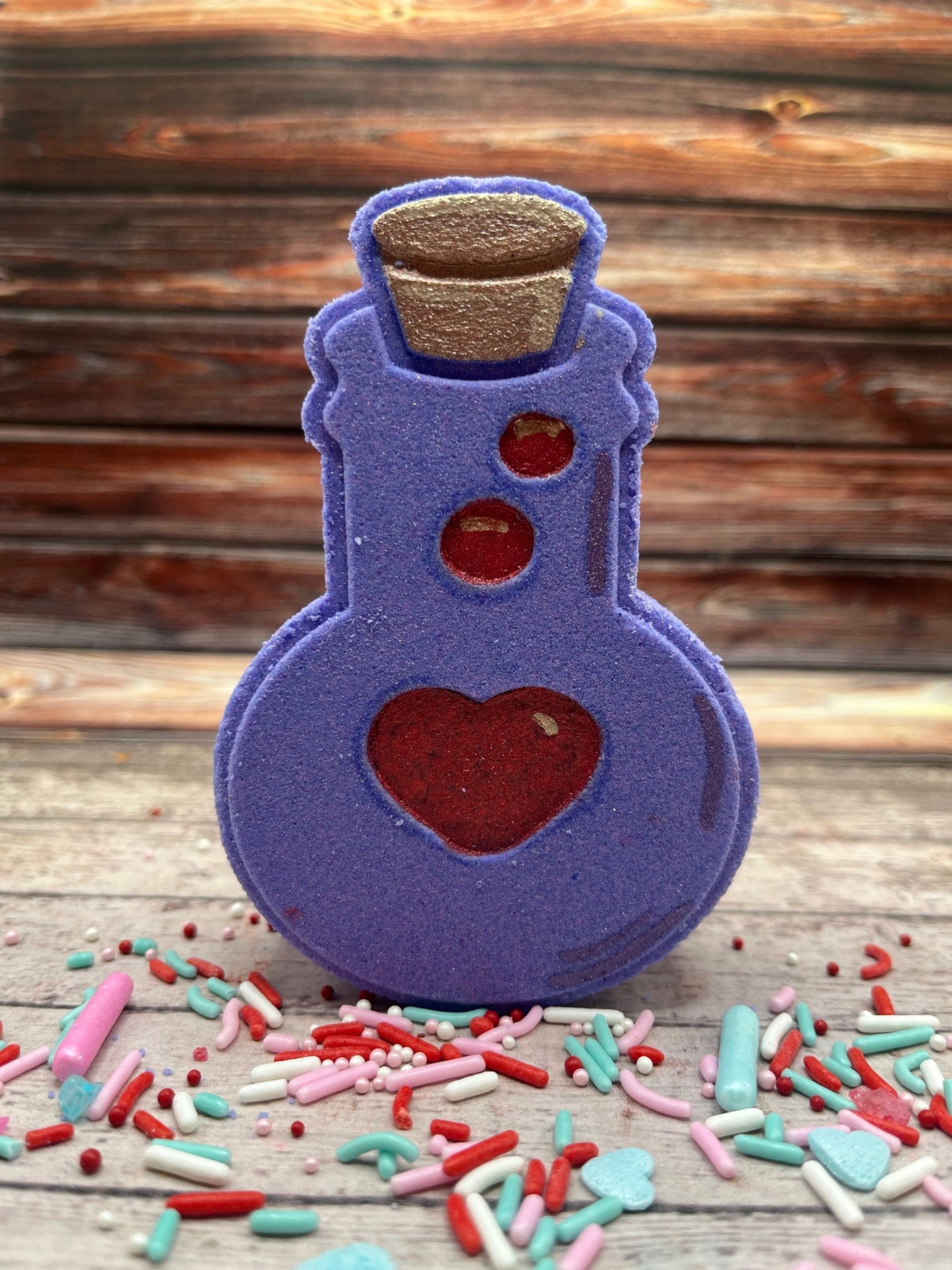 Purple potion bottle shaped bath bomb. Hand-painted red heart and bubbles along with tan cork. Displayed on rustic wooden background and pastel pink and blue sprinkles.