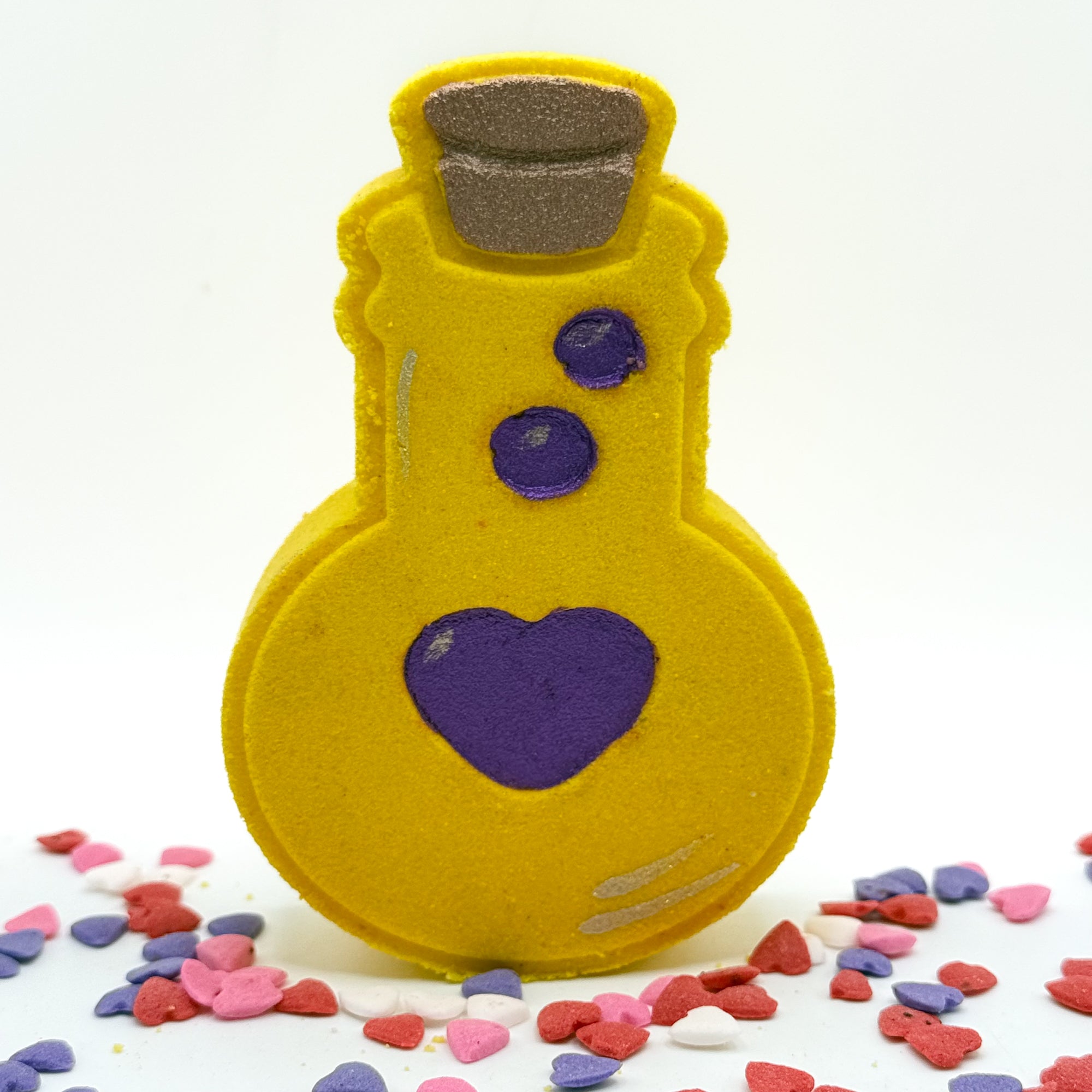 Purple potion bottle shaped bath bomb. Hand-painted red heart and bubbles along with tan cork. Displayed on rustic wooden background and pastel pink and blue sprinkles.