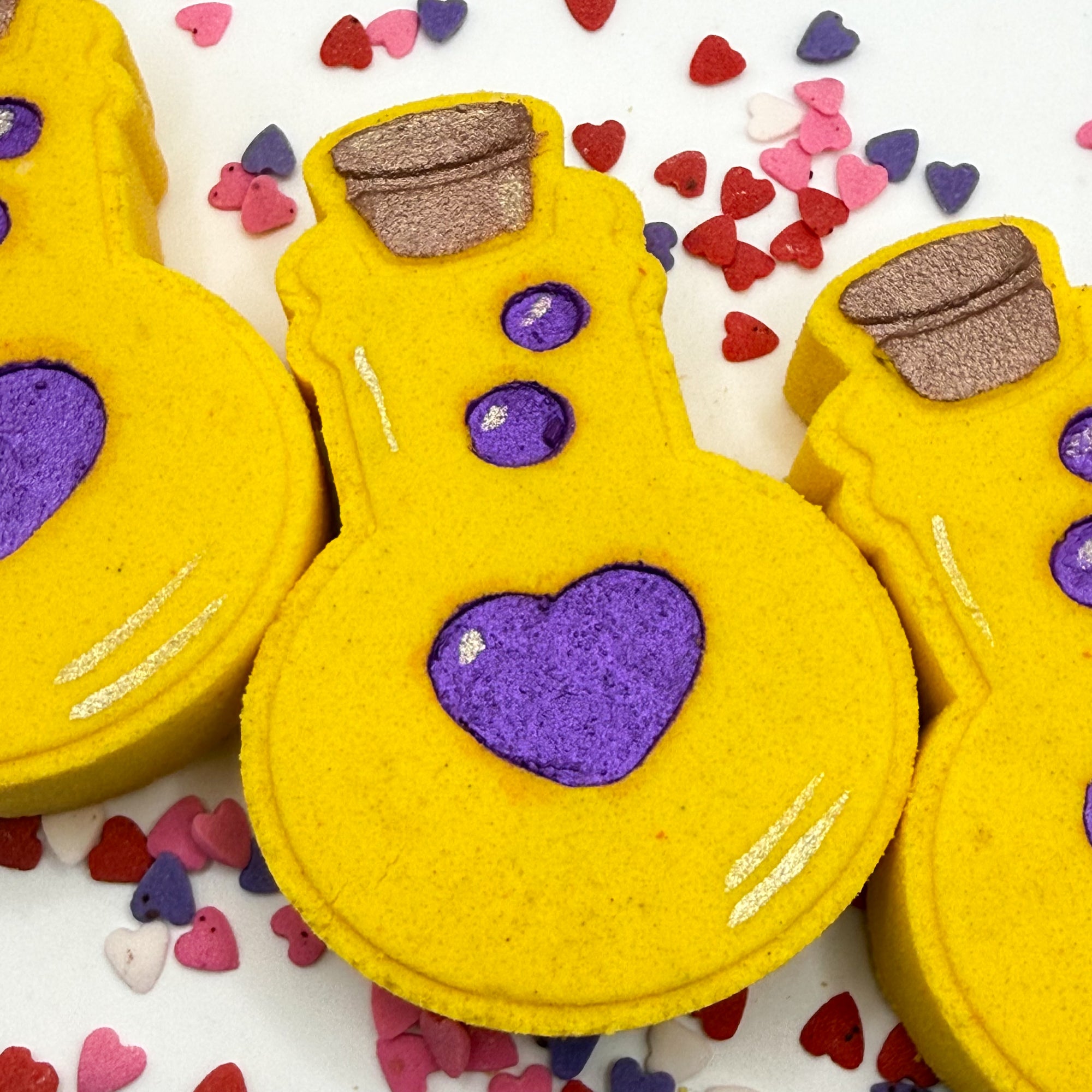 Hand painted yellow love potion bath bomb with purple hearts on a white background with colorful heart-shaped candies.