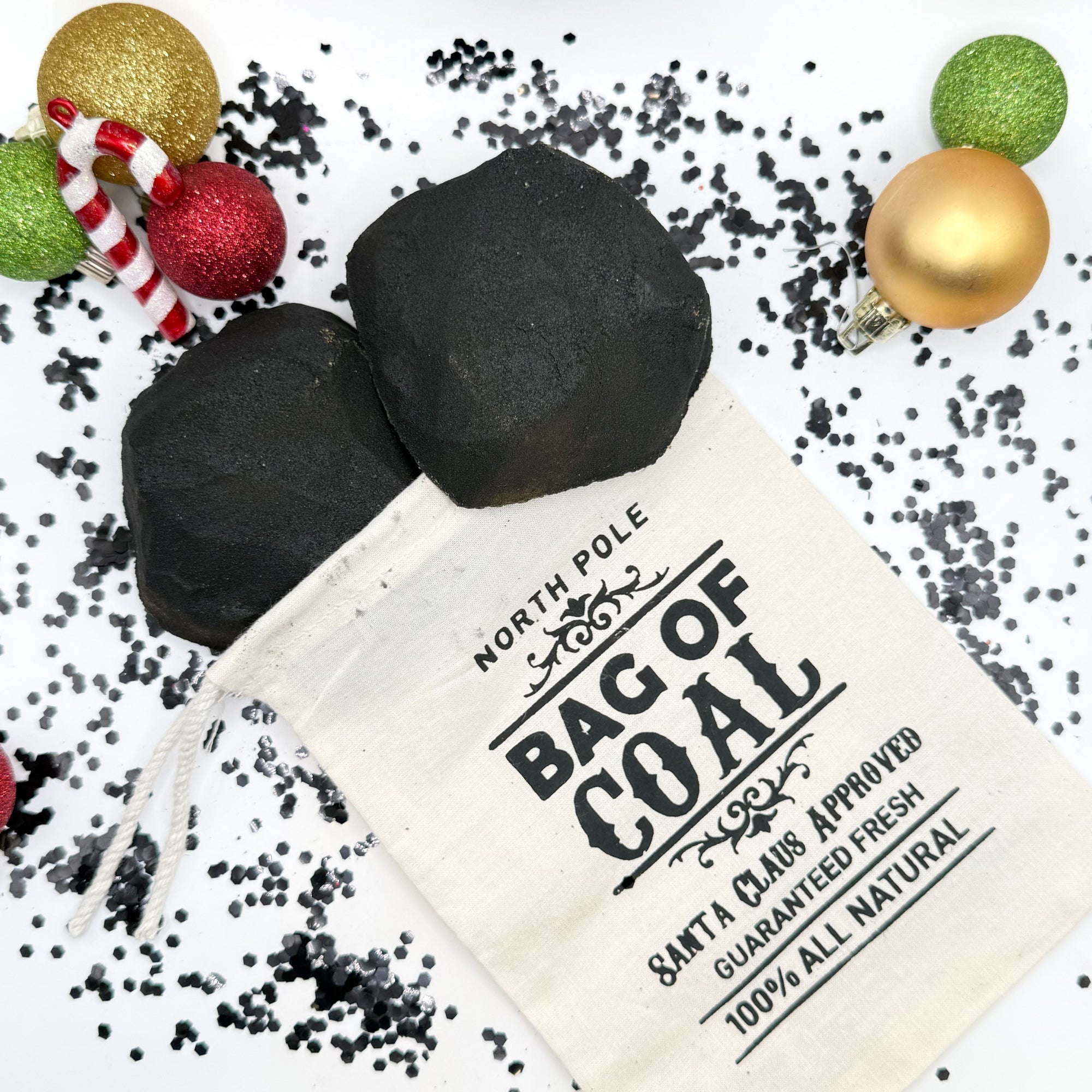 Two black lump of coal bath bombs with a tan 'Bag of Coal' pouch on a festive background with Christmas ornaments. Handmade by Rusted Acre Soap Co.