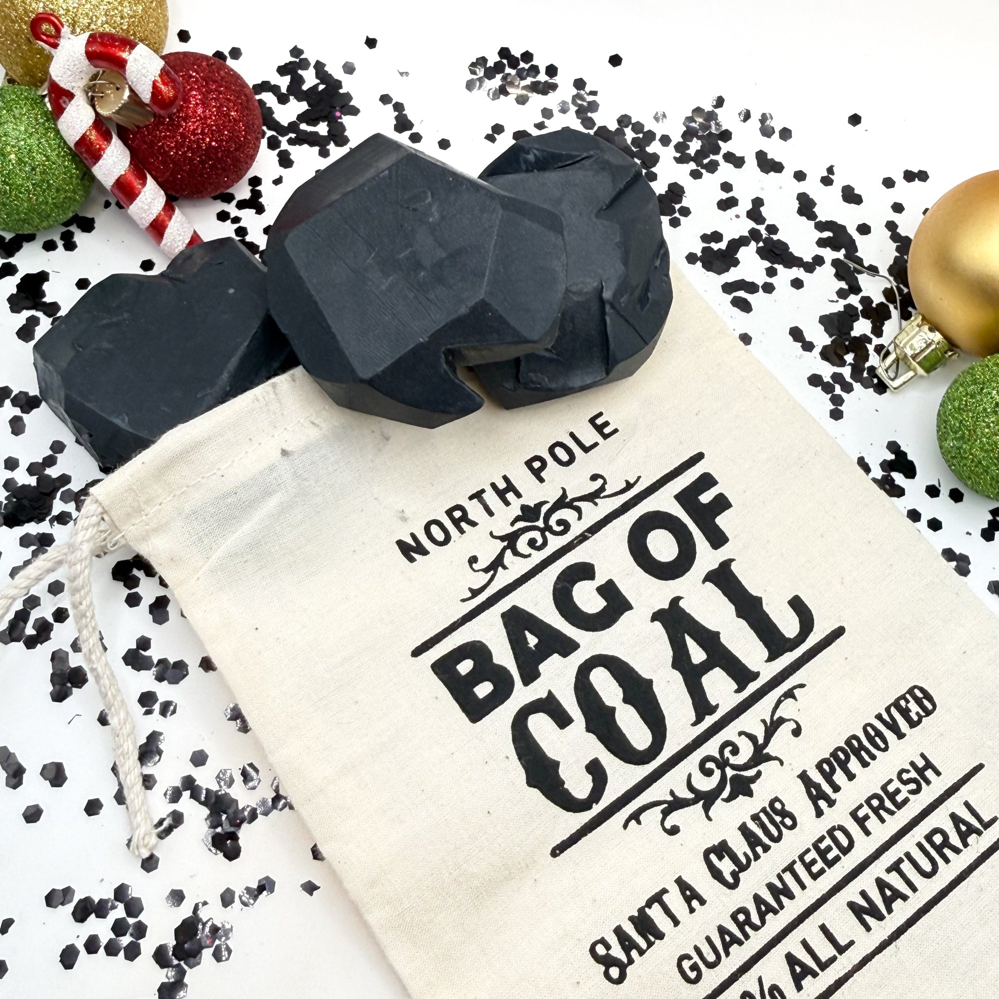 Three lump of coal soaps gag gift in a tan canvas bag on a festive background with ornaments.