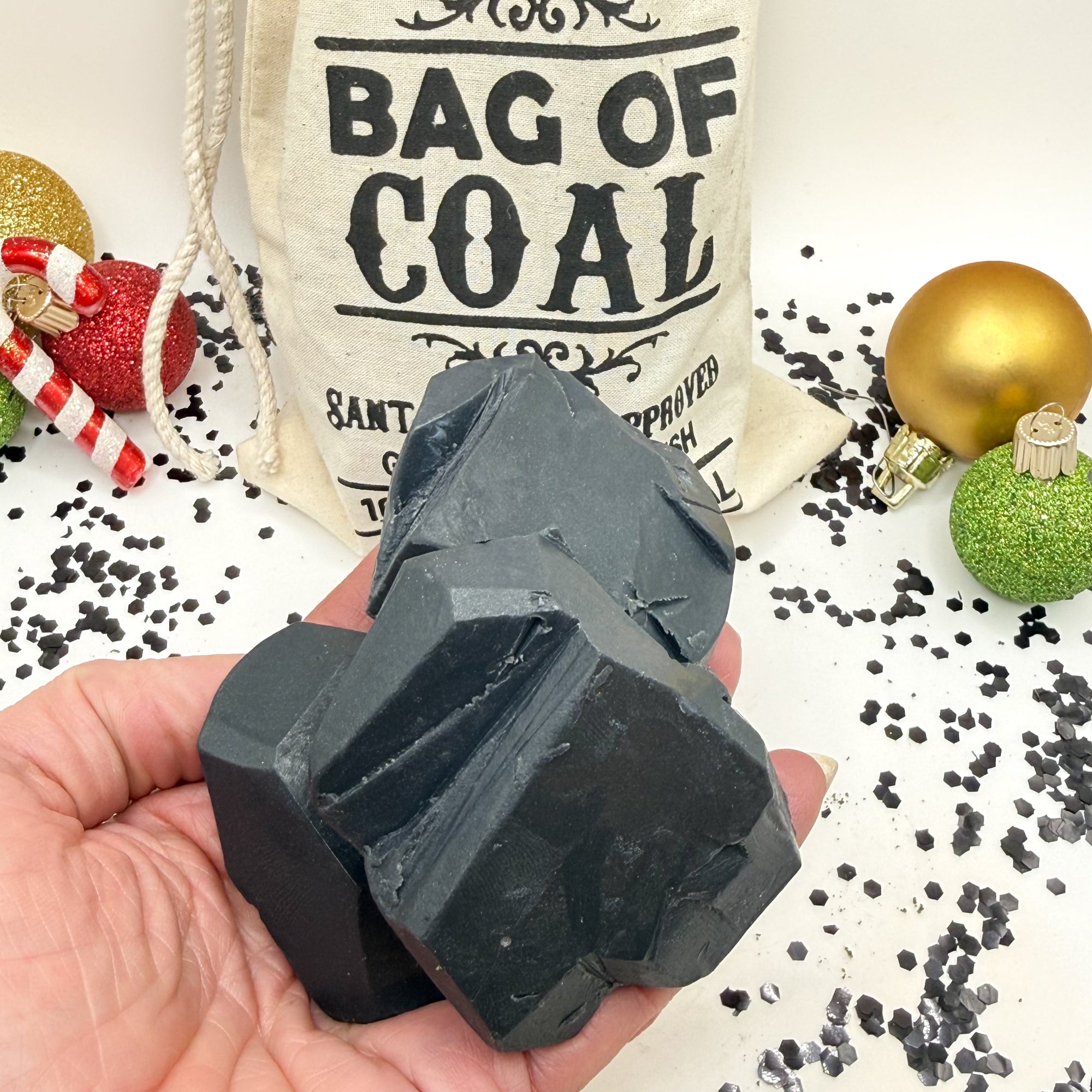 Hand holding three lump of coal soaps with a tan canvas 'Bag of Coal' sign and Christmas ornaments in the background from Rusted Acre Soap Co.