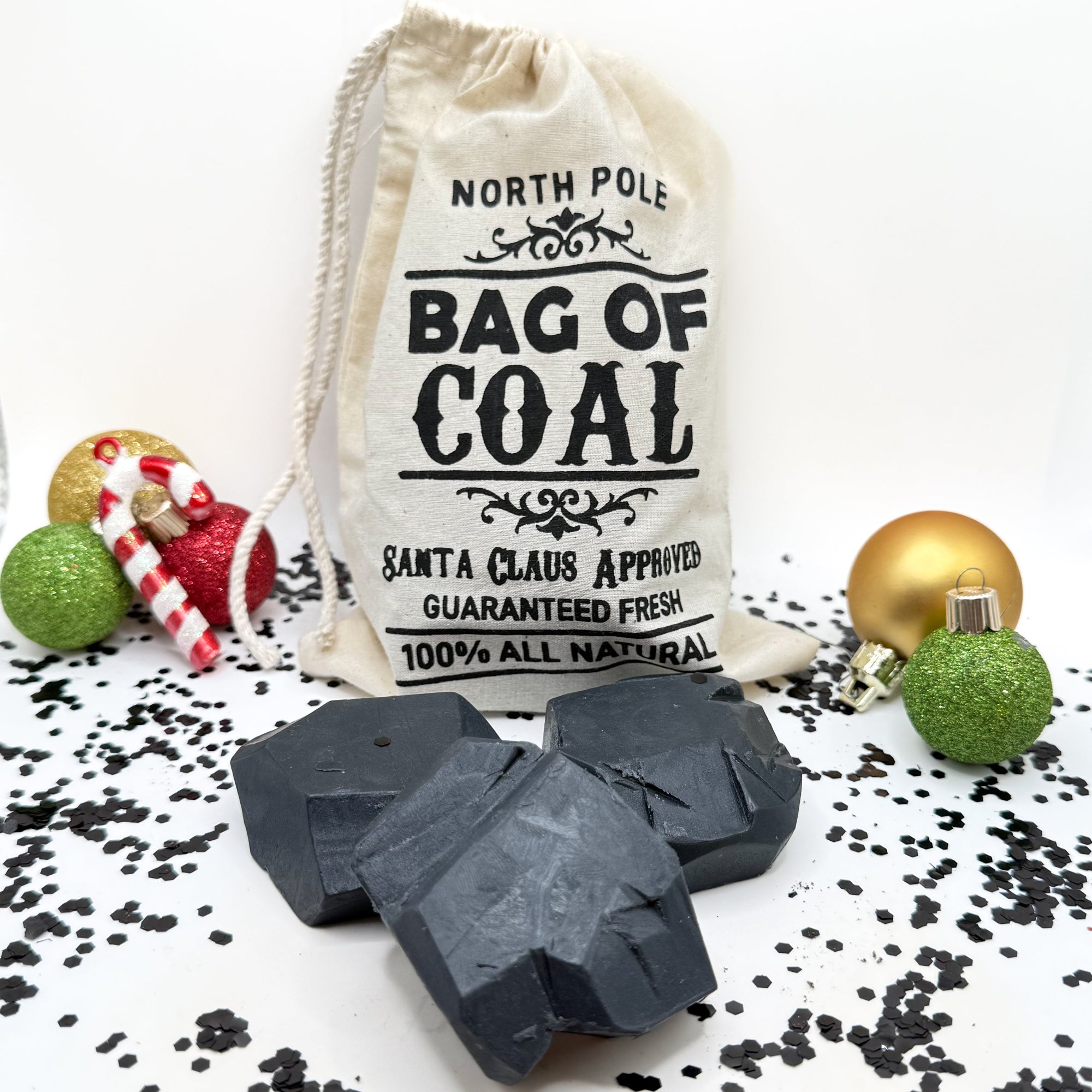 Three black lump of coal shaped soaps with a tan canvas "Bag of Coal" for Christmas on a festive background. Handmade by Rusted Acre Soap Co.