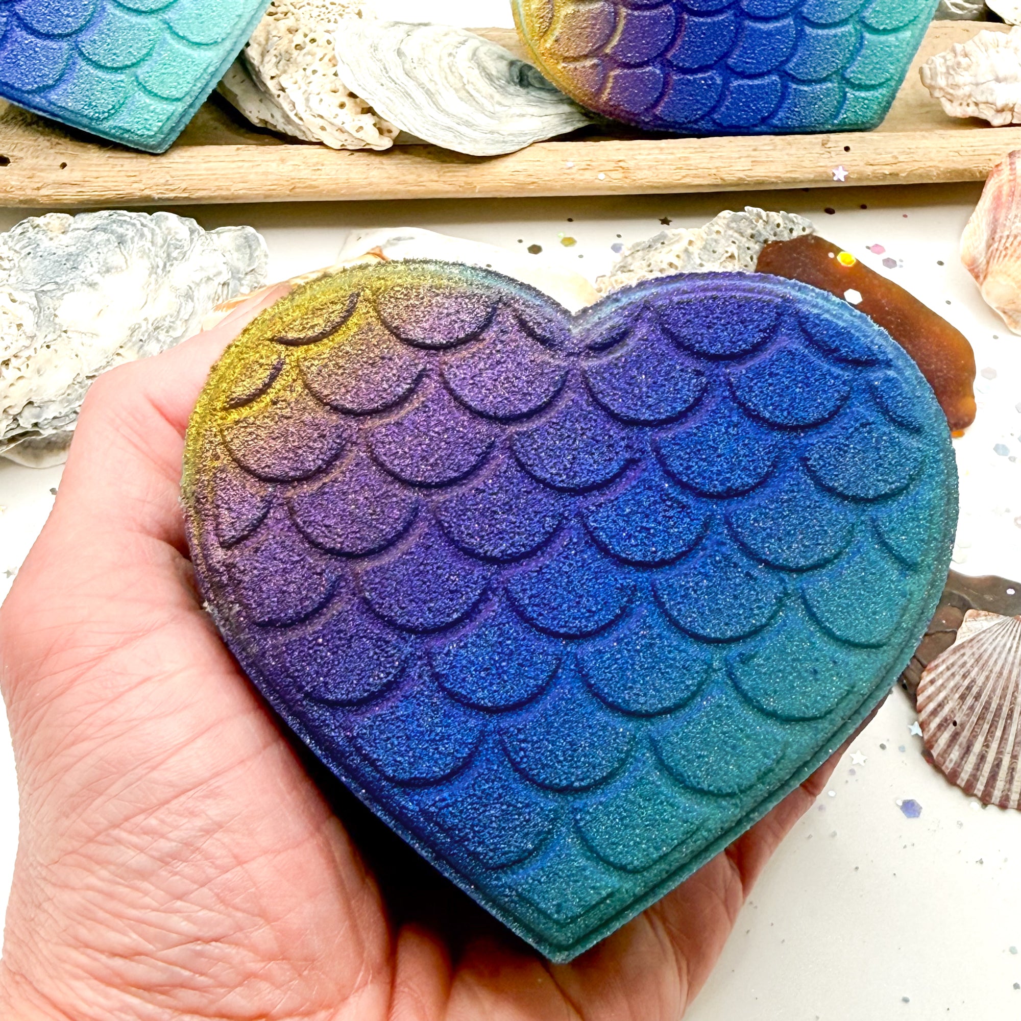 Heart shaped bath bomb with mermaid scale pattern held by a hand, with seashells in the background.