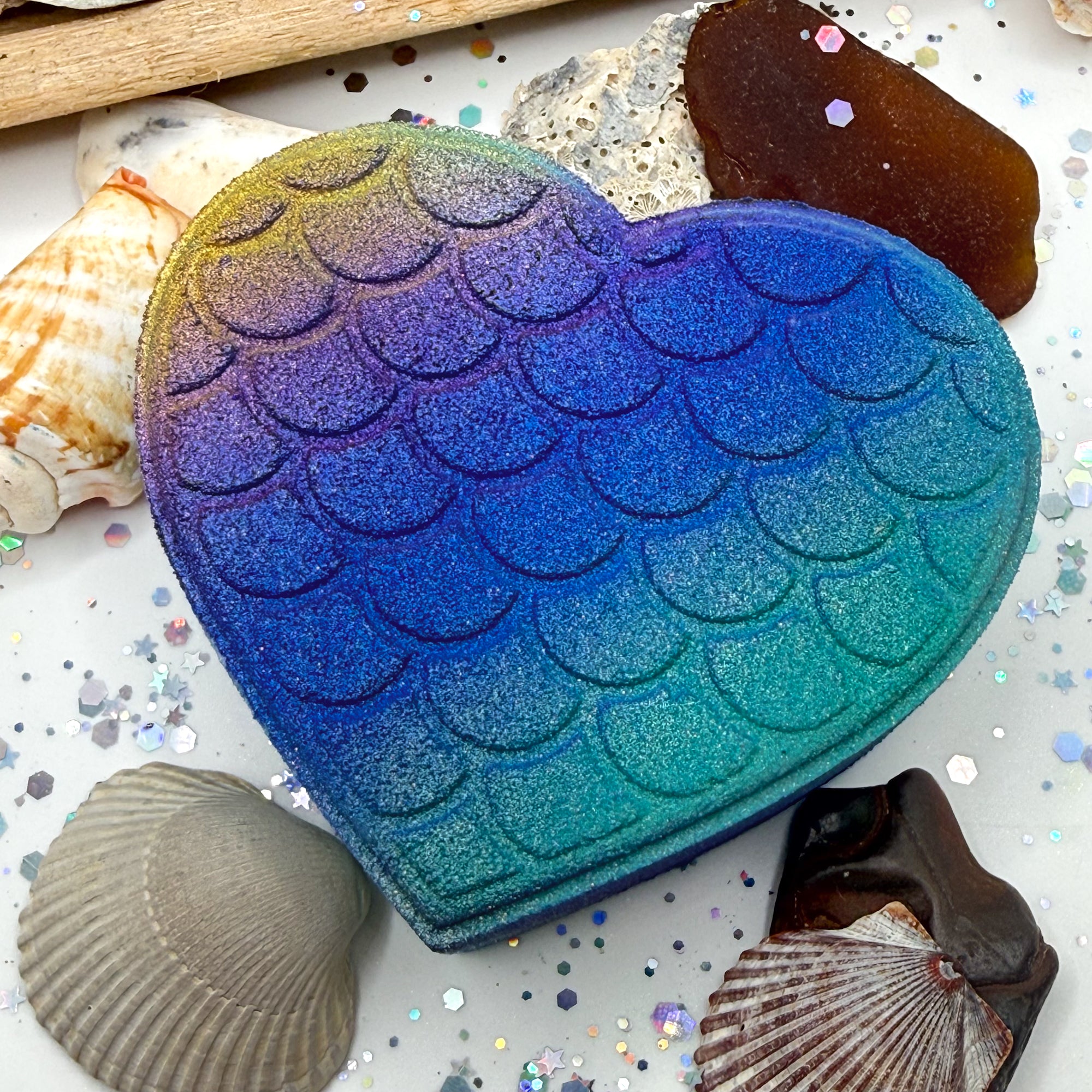 Teal, heart shaped bath bomb with mermaid scales. Purple to teal airbrushed gradient. Gold accent on the left side. Displayed on a rustic wooden background.
