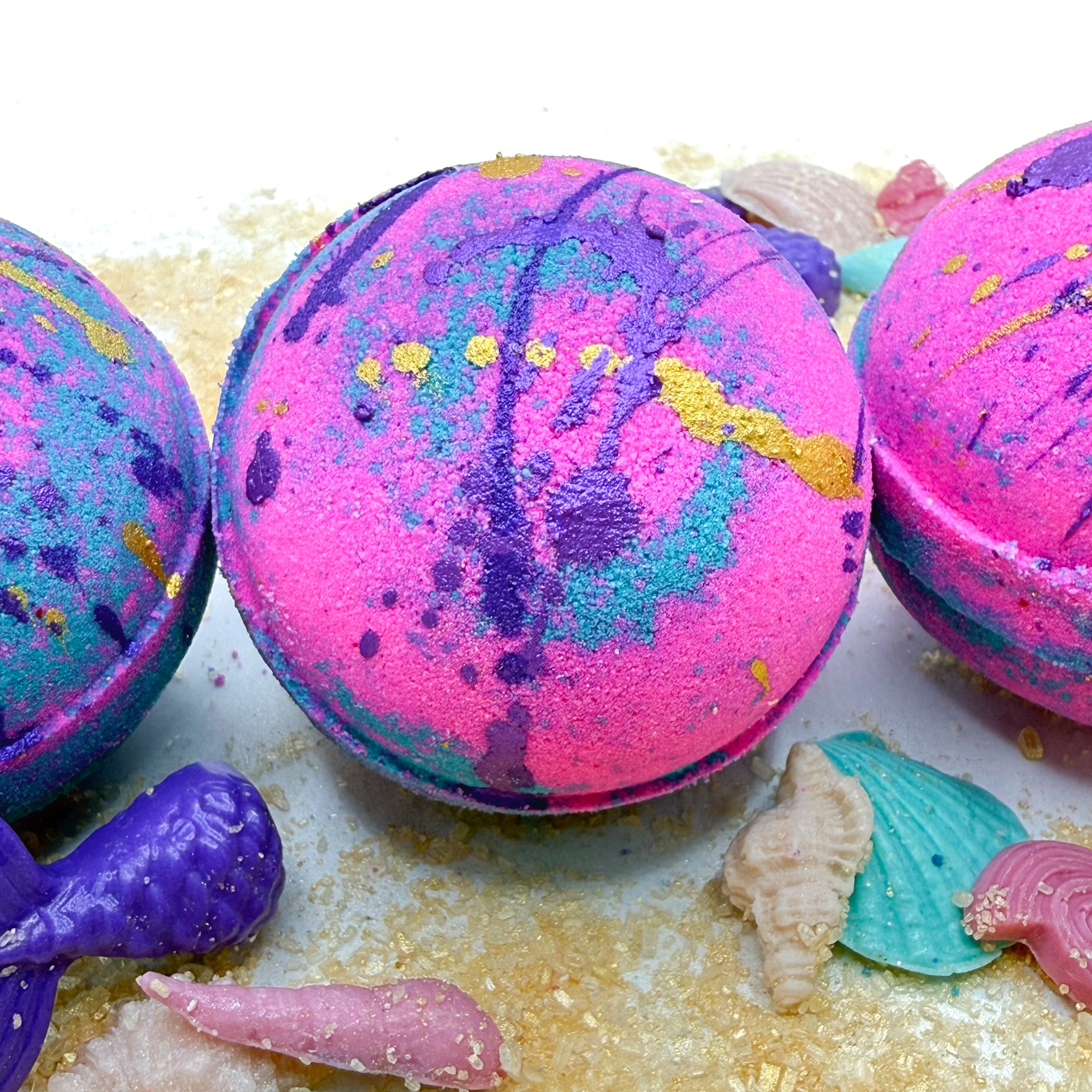 Teal and pink bath bombs with a seashell and starfish decorations on a sandy background.