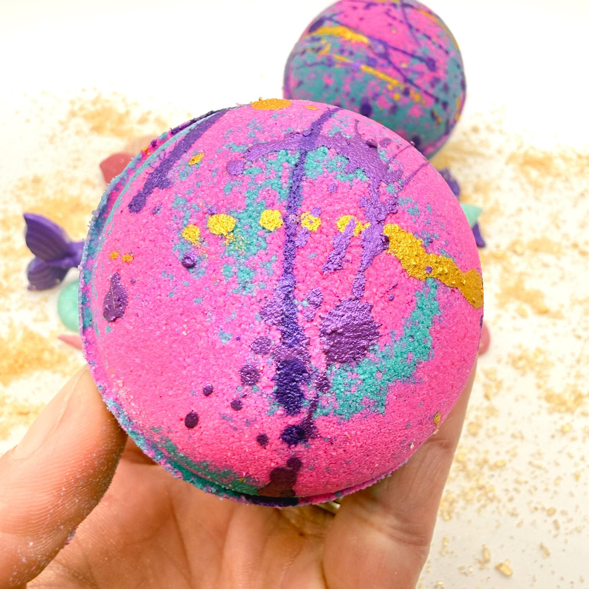 Pink and teal bath bomb held in a hand with a blurred beach themed background.