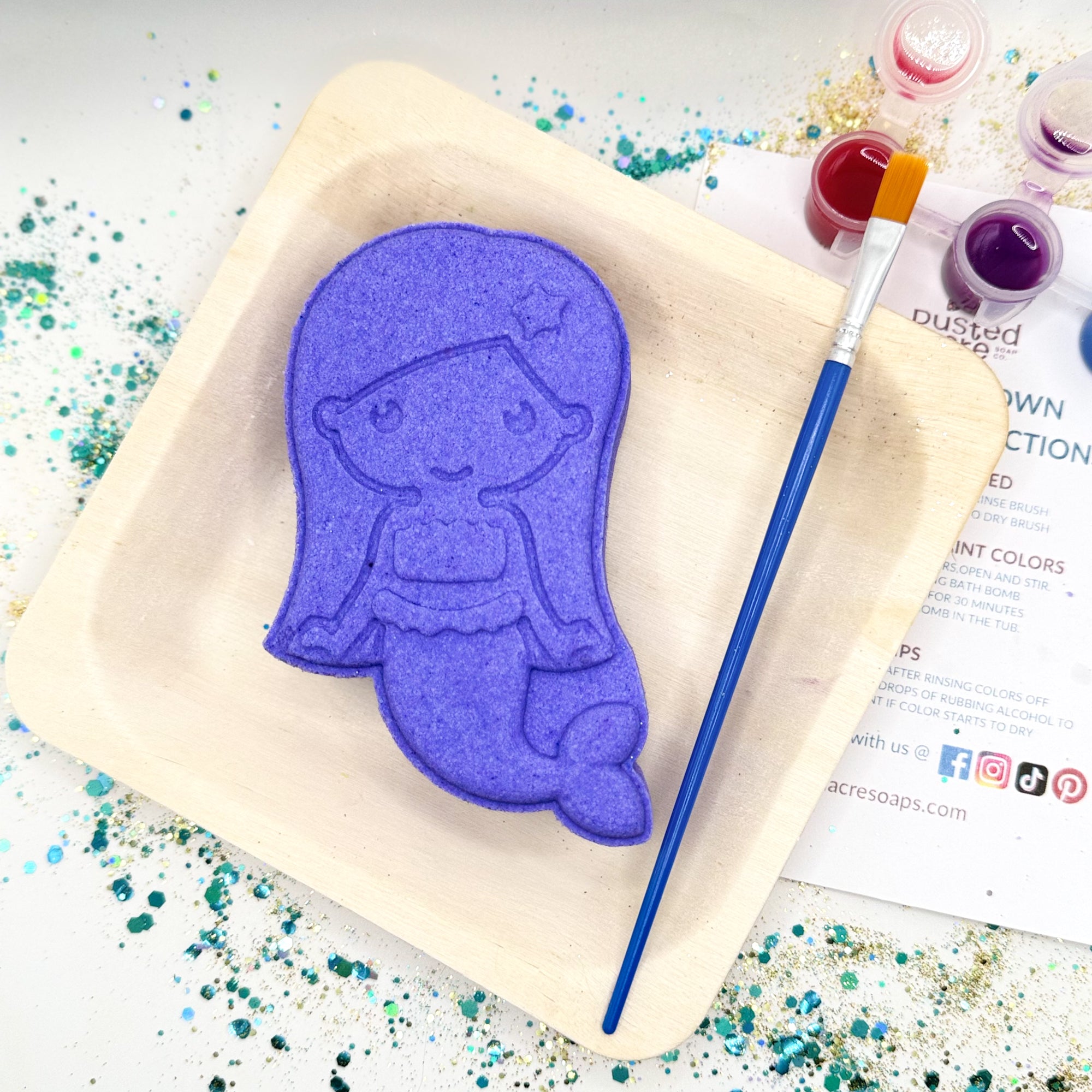 Purple mermaid paint your bath bomb kit on a wood plate with a paintbrush and colorful paint splashes.