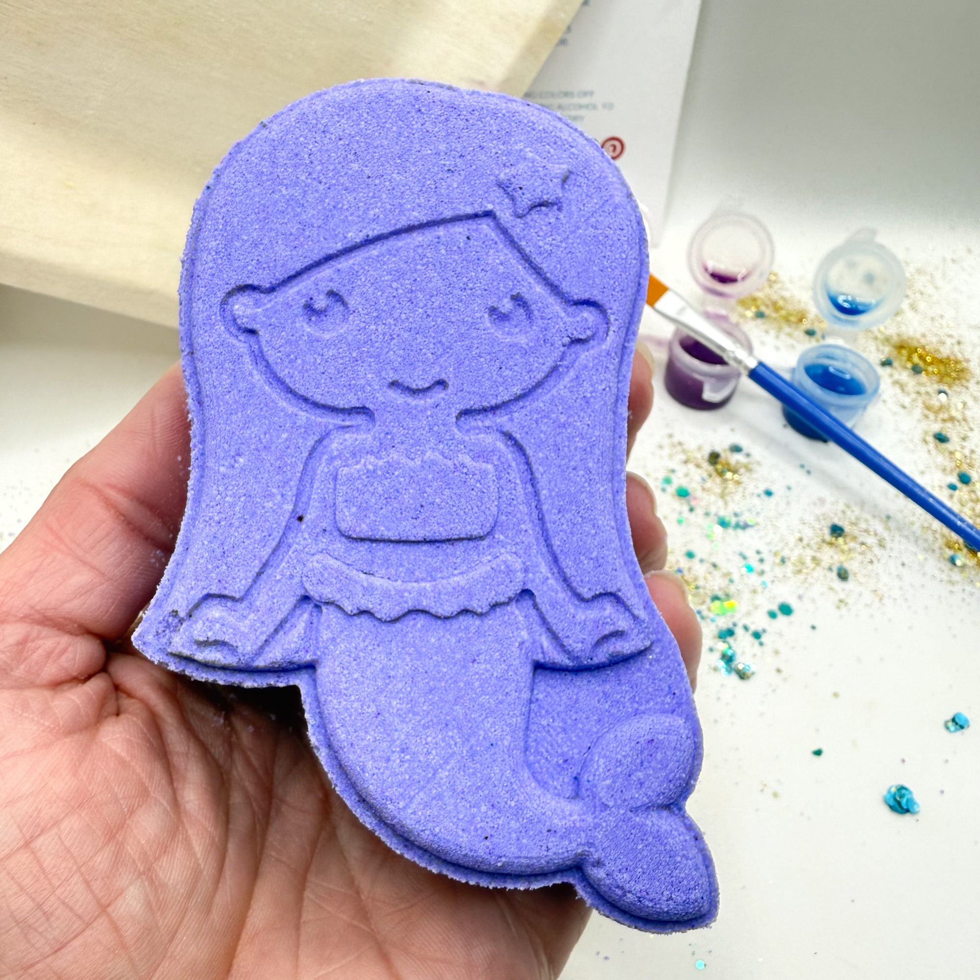 Purple paint your own mermaid bath bomb held by a hand, with painting materials in the background.