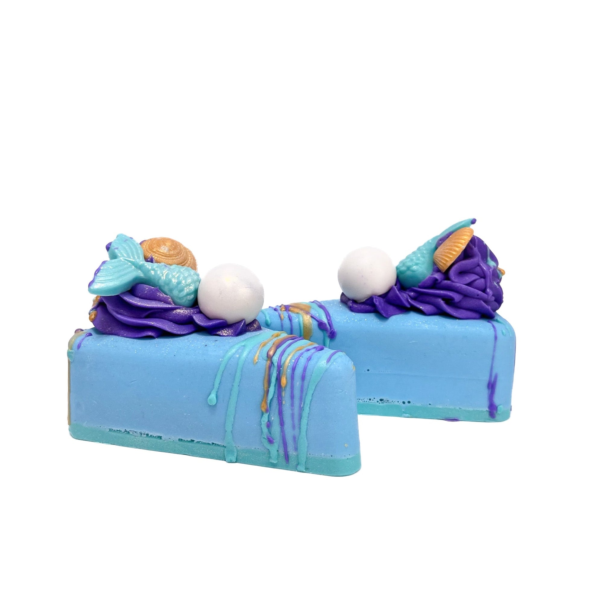 Two blue pie slice shaped bars of soap. Topped with a puff of soap, a mermaid teal mermaid tail soap embed, a white pearl soap embed, drizzled with teal, purple, and gold soap.