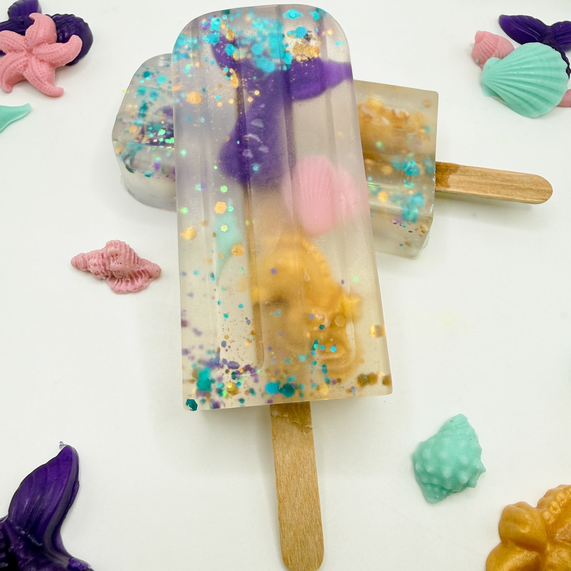 Two hand poured popsicle soaps with mermaid themed soap embeds inside clear soap on popsicle sticks.