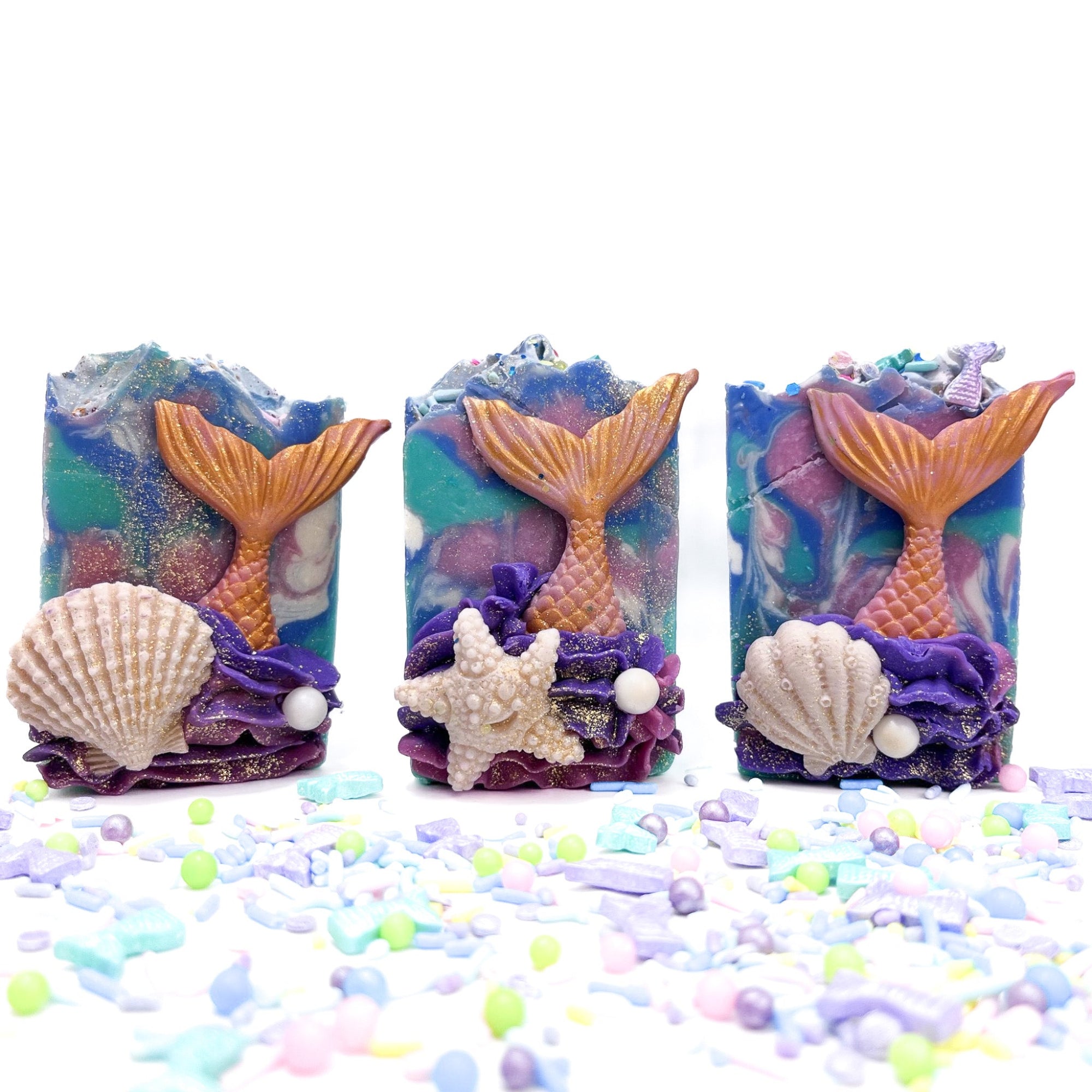 Three bars of Rusted Acre Soap Company's Mermaid Tail handmade soap. Two have white clam shell embeds one has a starfish.