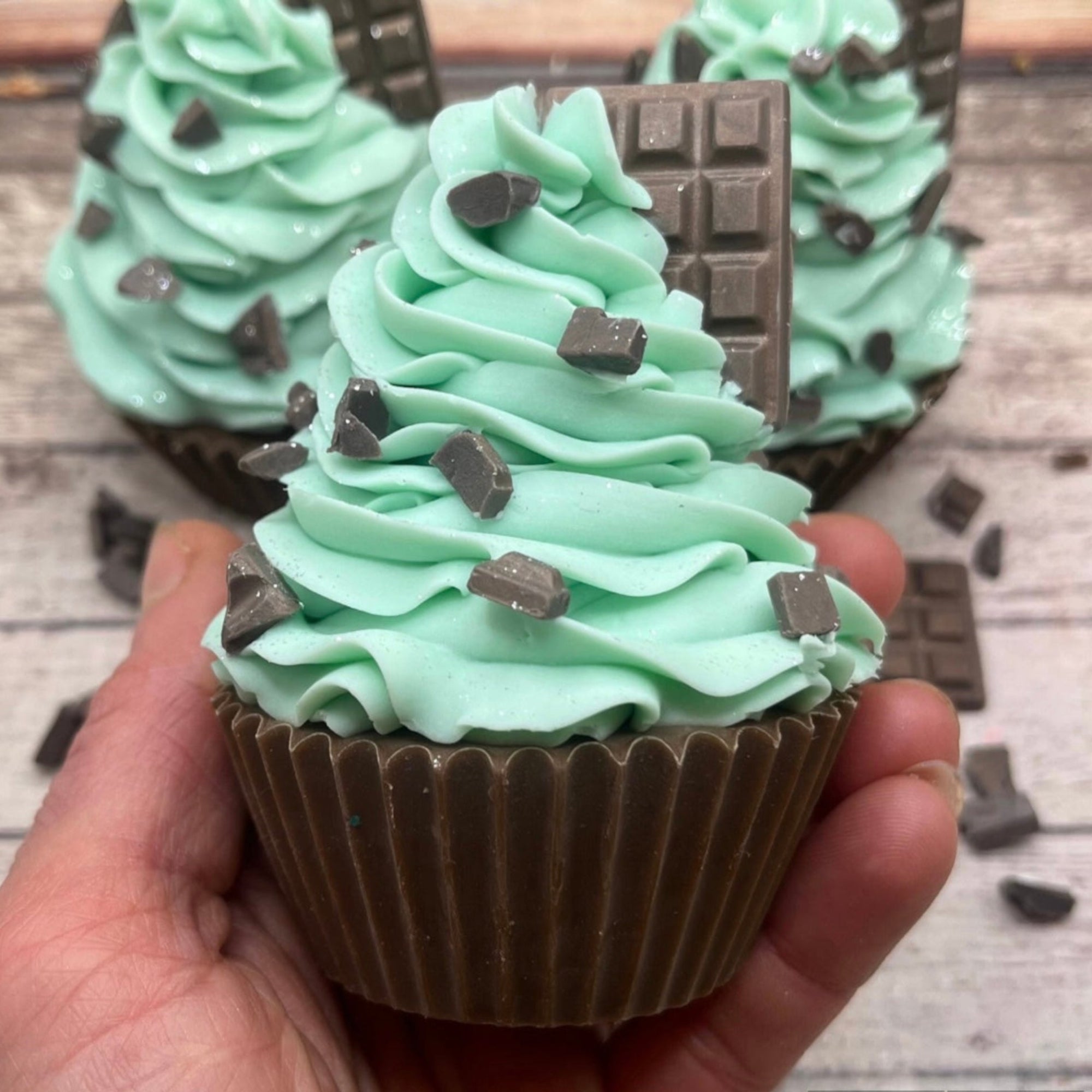 One hand piped and decorated mint chocolate chip cupcake soap being held in a hand. Soap is decorated with soap “chocolate chips” and a little soap chocolate bar.