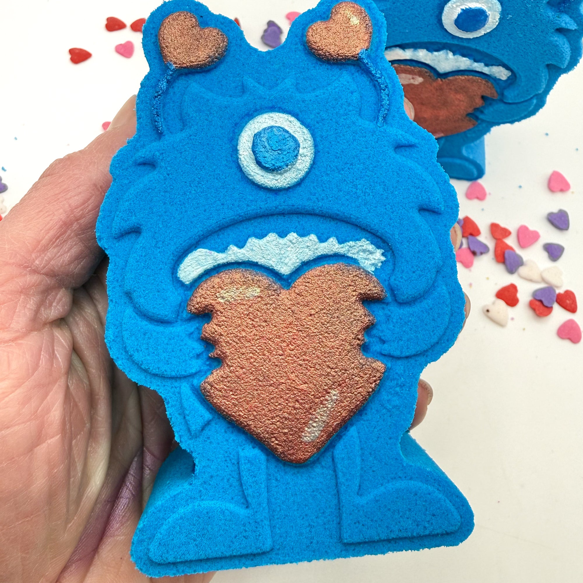 Hand painted blue monster shaped bath bomb being held in a hand with heart shape candies around it.