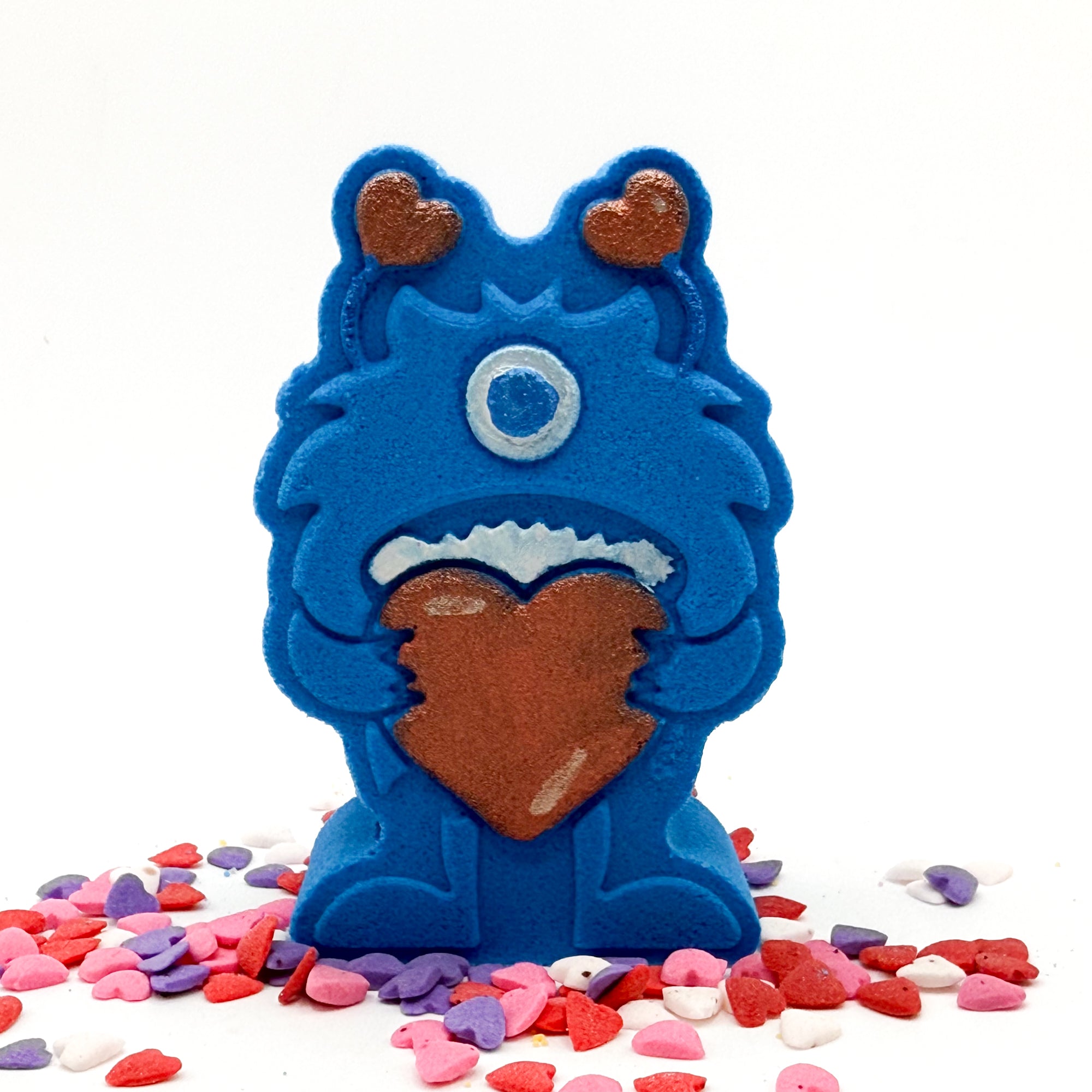 Hand painted blue monster shaped bath bomb holding a red hart with red hart antennas with heart shape candies around it.