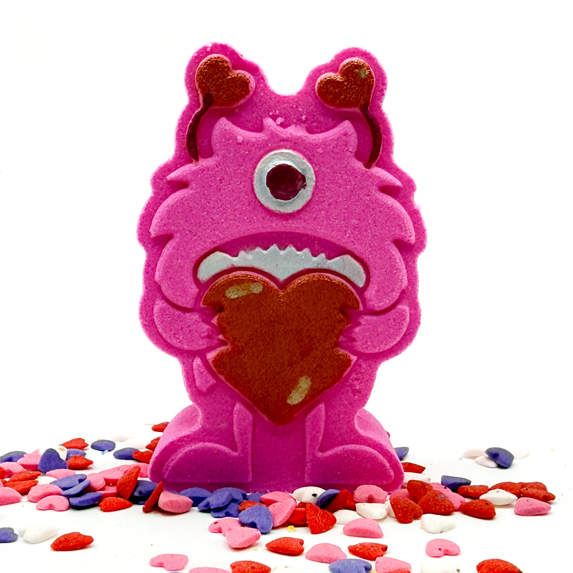 Hand painted Pink monster shaped bath bomb holding a red hart with red hart antennas with heart shape candies around it.