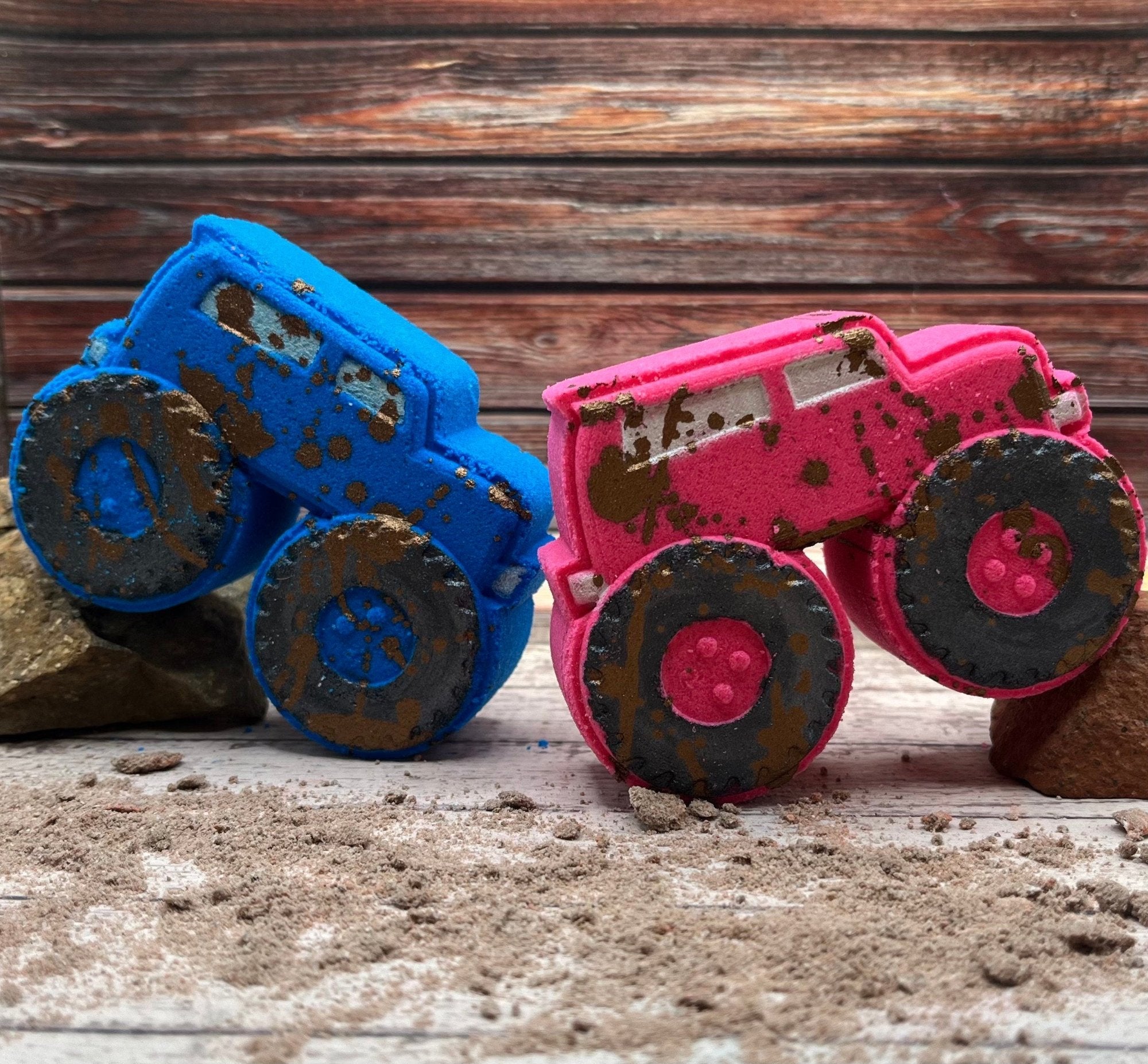 Two S-U-V style trucks. One pink and one blue hand-painted silver windows and black tires, and splattered with mud colored mica. They are arranged like they are climbing boulders.