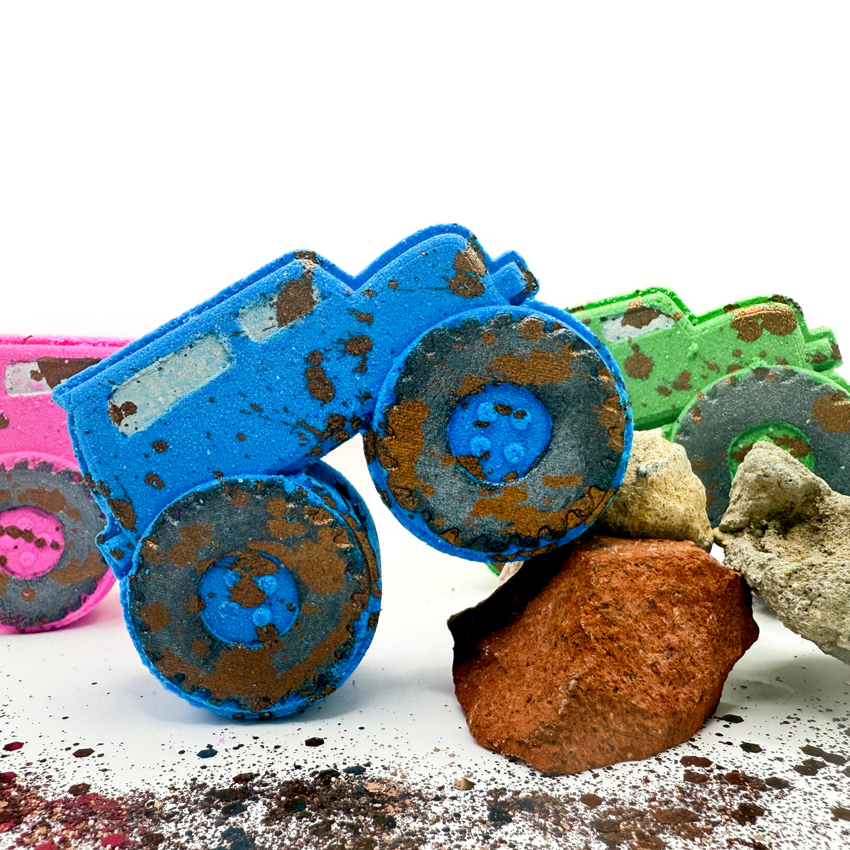Three monster truck shaped bath bombs in blue, green, and pink,with hand painted mud splatter and placed on rocks on a white surface against a white background.
