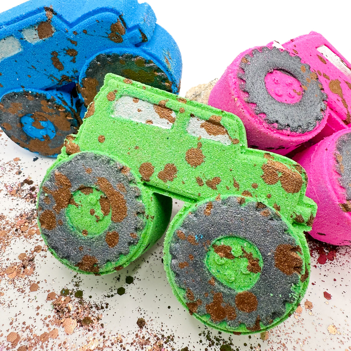 Three truck shaped bath bombs in blue, green, and pink, decorated with glittery black wheels and brown glitter mud splatters, arranged on a white surface.