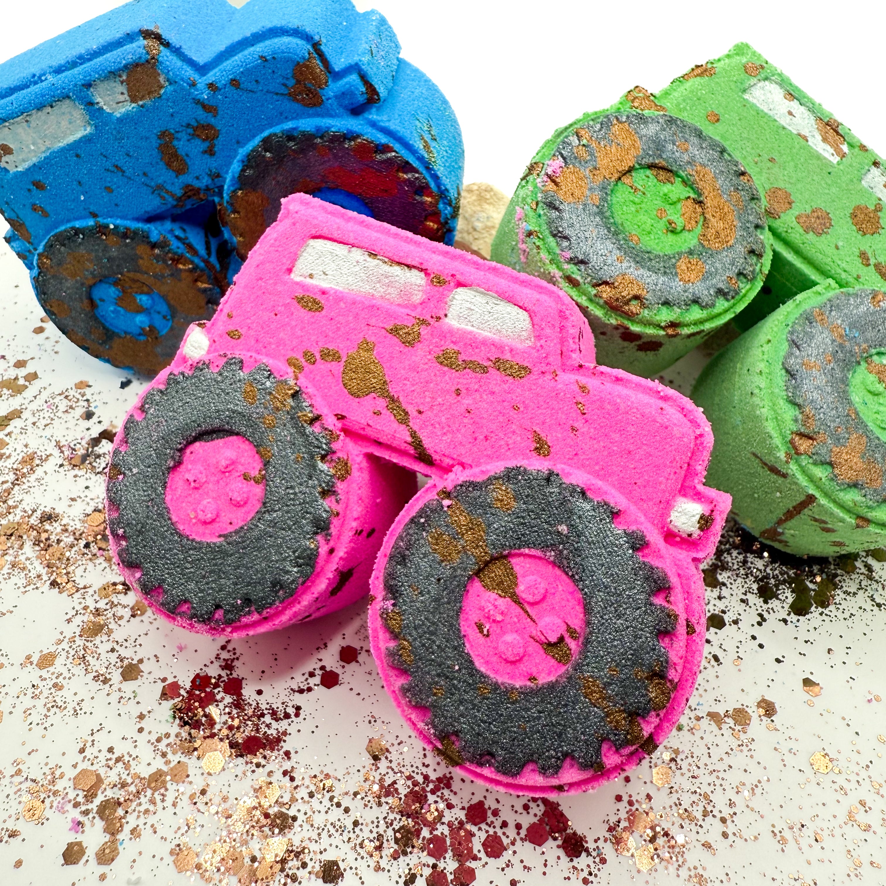 Three colorful monster truck shaped bath bombs or soaps arranged on a white surface with glitter. The front truck is bright pink with black treaded tires and brown splatter, while the other two are blue and green with similar treaded details.