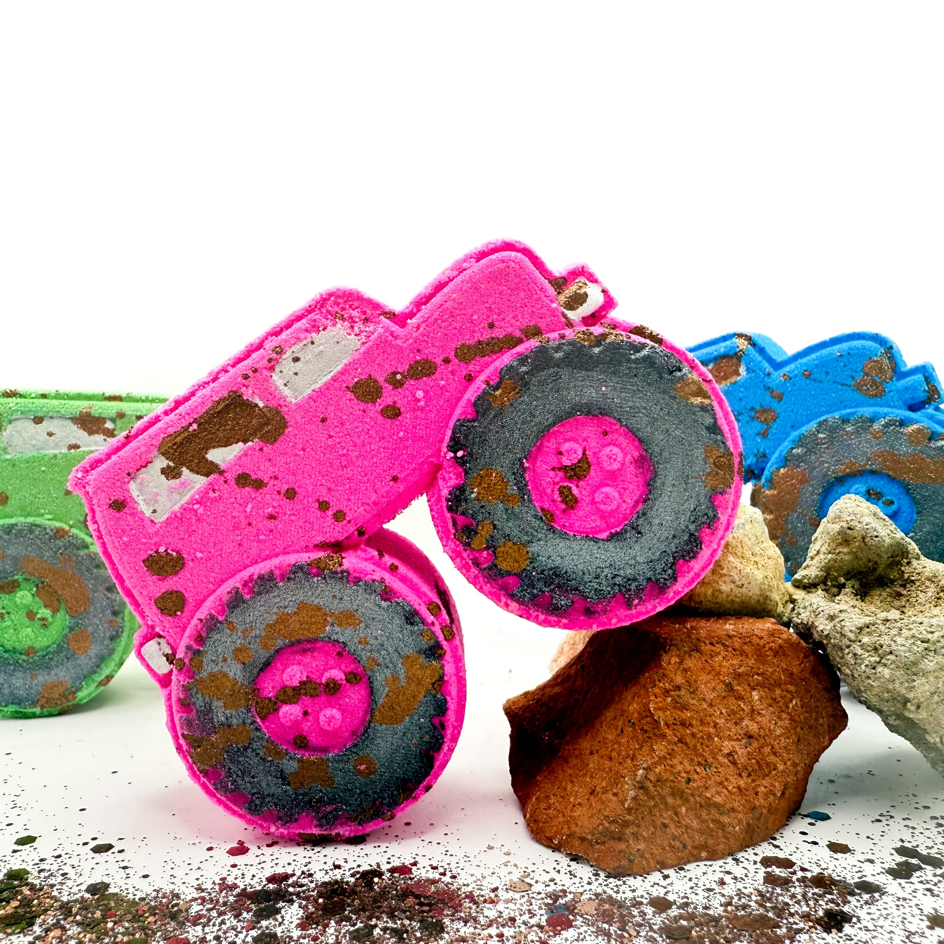 A bright pink monster truck with hand painted black wheels and brown mud like speckling, posed on white with rocks and other colorful bath bombs partially visible in the background.