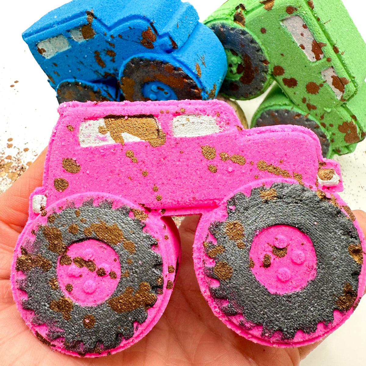 A close up of a hand holding a pink monster truck bath bomb with hand painted brown mud-splattered and black tires. Two more monster truck bath bombs, one blue and one green, are visible in the background on a white surface.