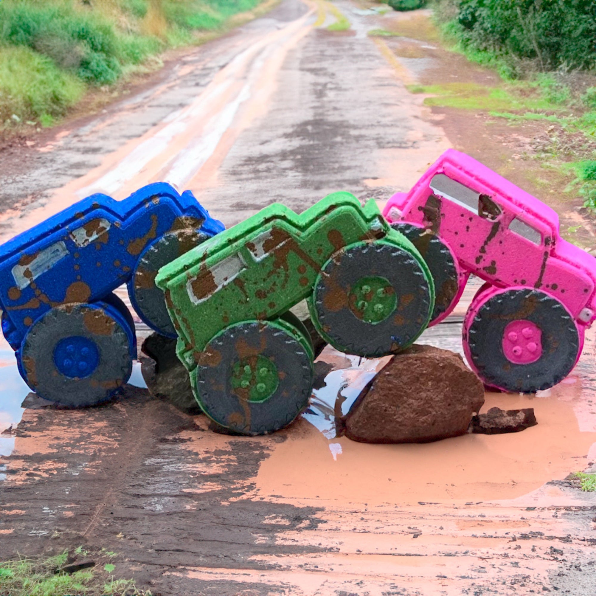 A group of three muddy monster truck bath bombs in pink, blue, and green, each with black wheels and muddy brown splashes, displayed together.