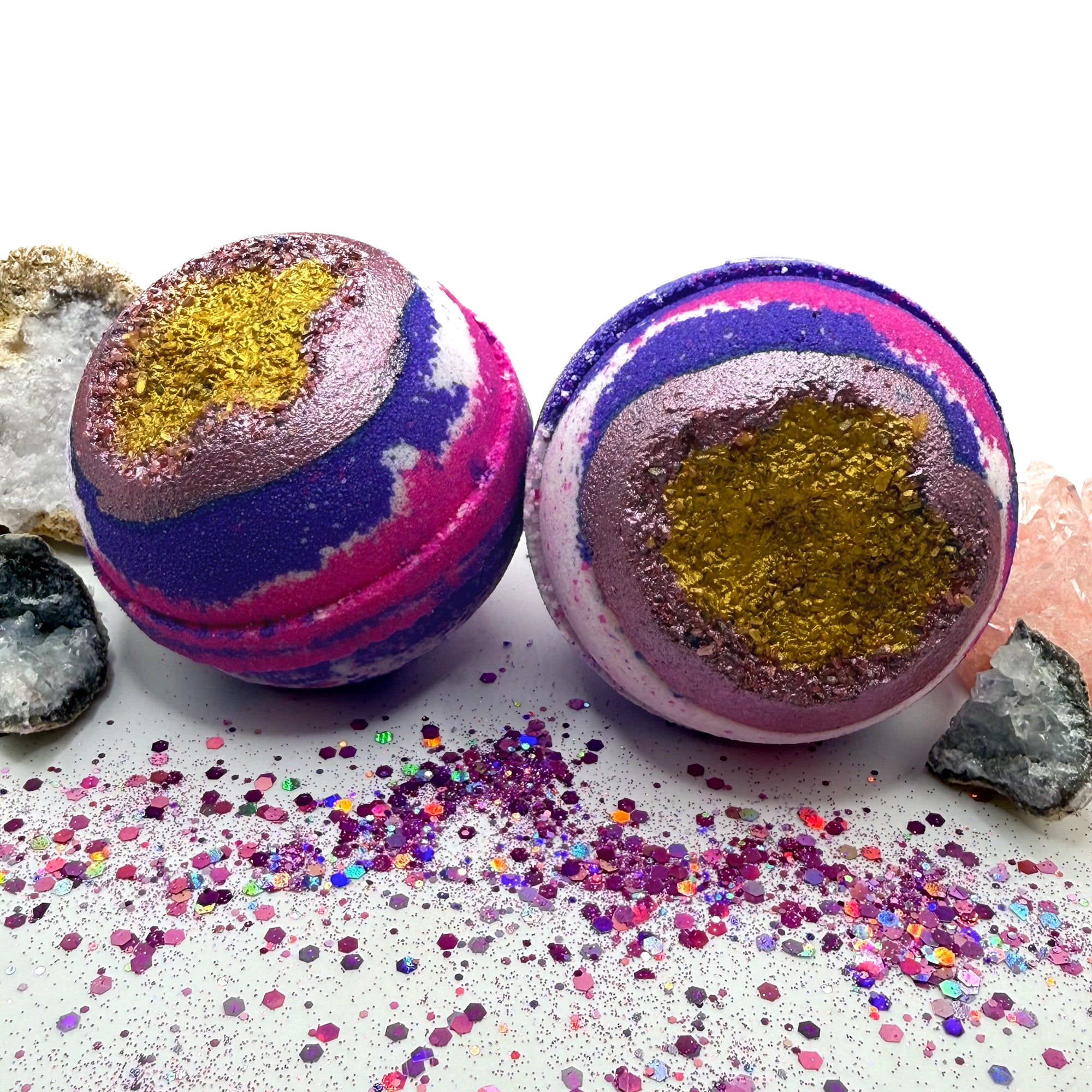 Two pink, purple and white bath bombs with a geode inspired center on a white surface with glitter scattered around.