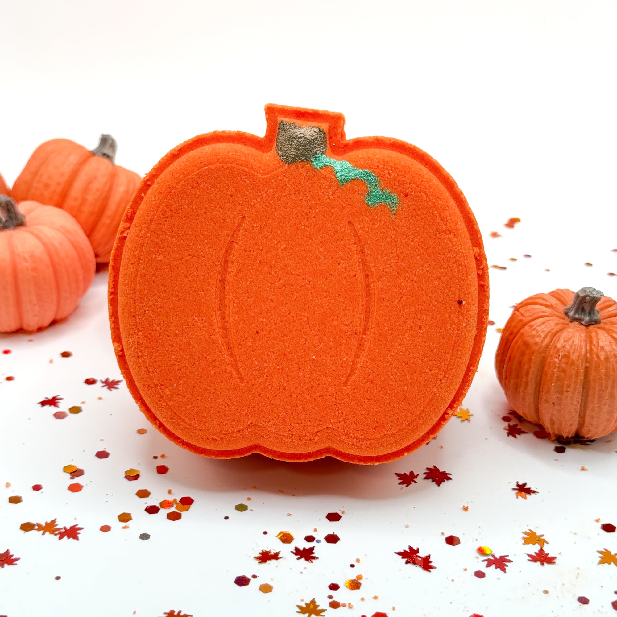An orange pumpkin-shaped bath bomb with hand painted details, displayed on a white surface with scattered autumn confetti and small decorative pumpkins in the background.