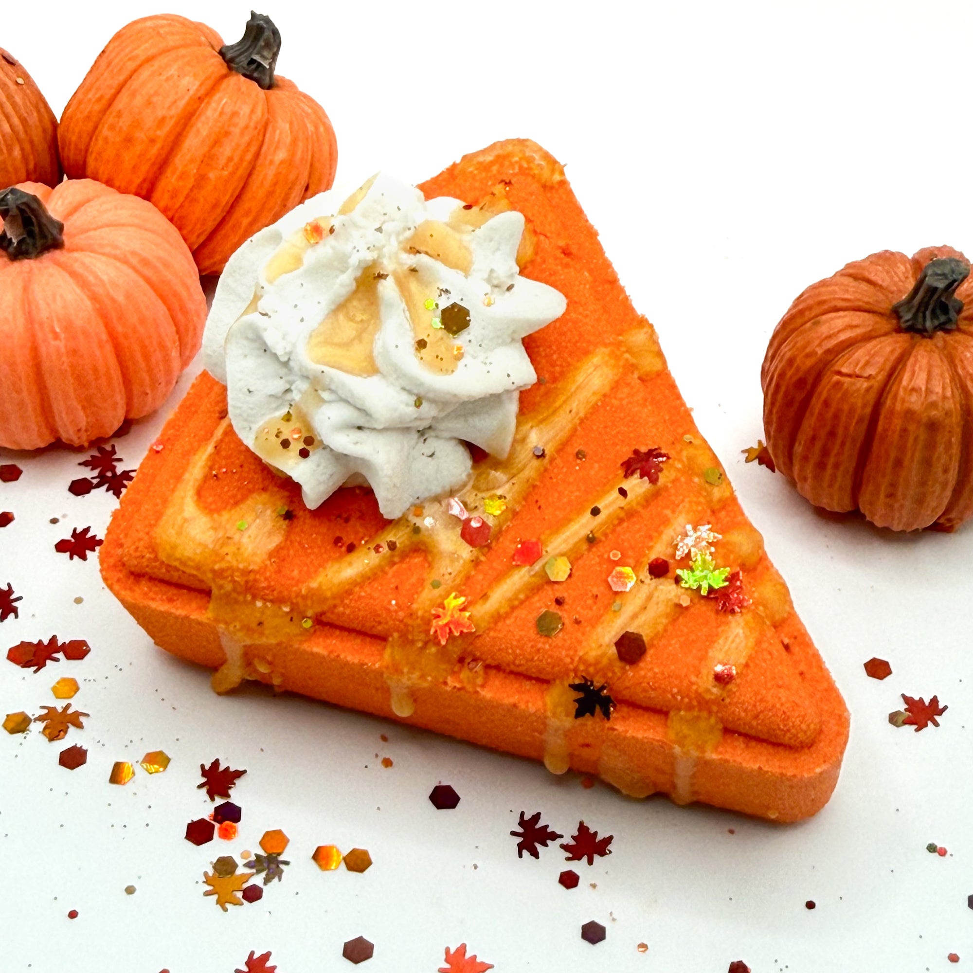 An orange pumpkin pie shaped bath bomb topped with white whipped cream, drizzled caramel-colored syrup, and sprinkled with glitter, set on a white background with decorative pumpkins and autumn leaves.