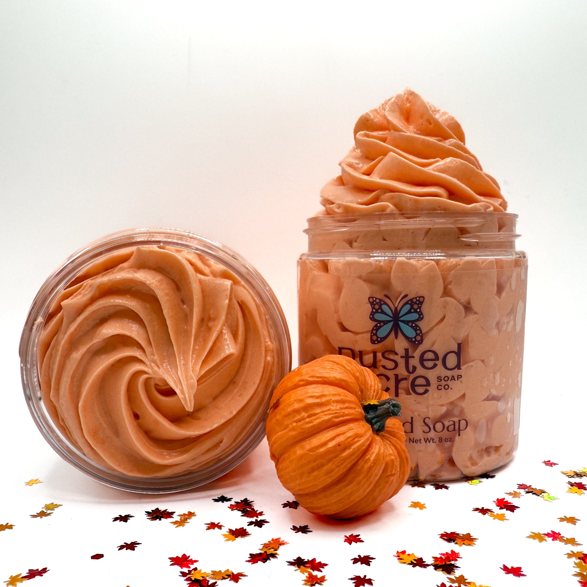 Two jars of orange pumpkin spicewhipped soap with a small pumpkin and fall leaf confetti on a white background.