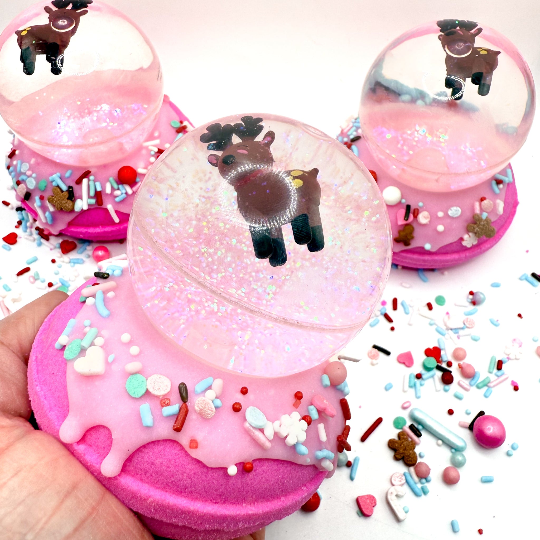 Pink donut-shaped bath bomb topped with a reindeer bouncy ball to make a snow globe shape in a hand with colorful sprinkles on a white background. Handmade by Rusted Acre Soap Co.