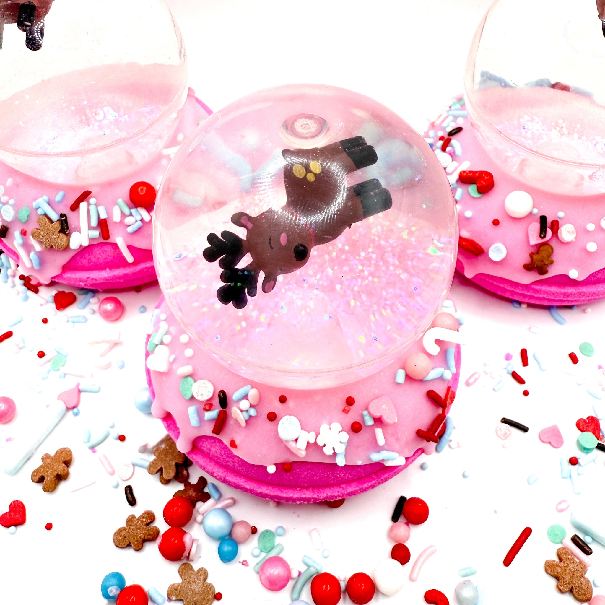 Three pink donut-shaped bath bombs topped with reindeer bouncy balls surrounded by colorful sprinkles and candies on a white background.