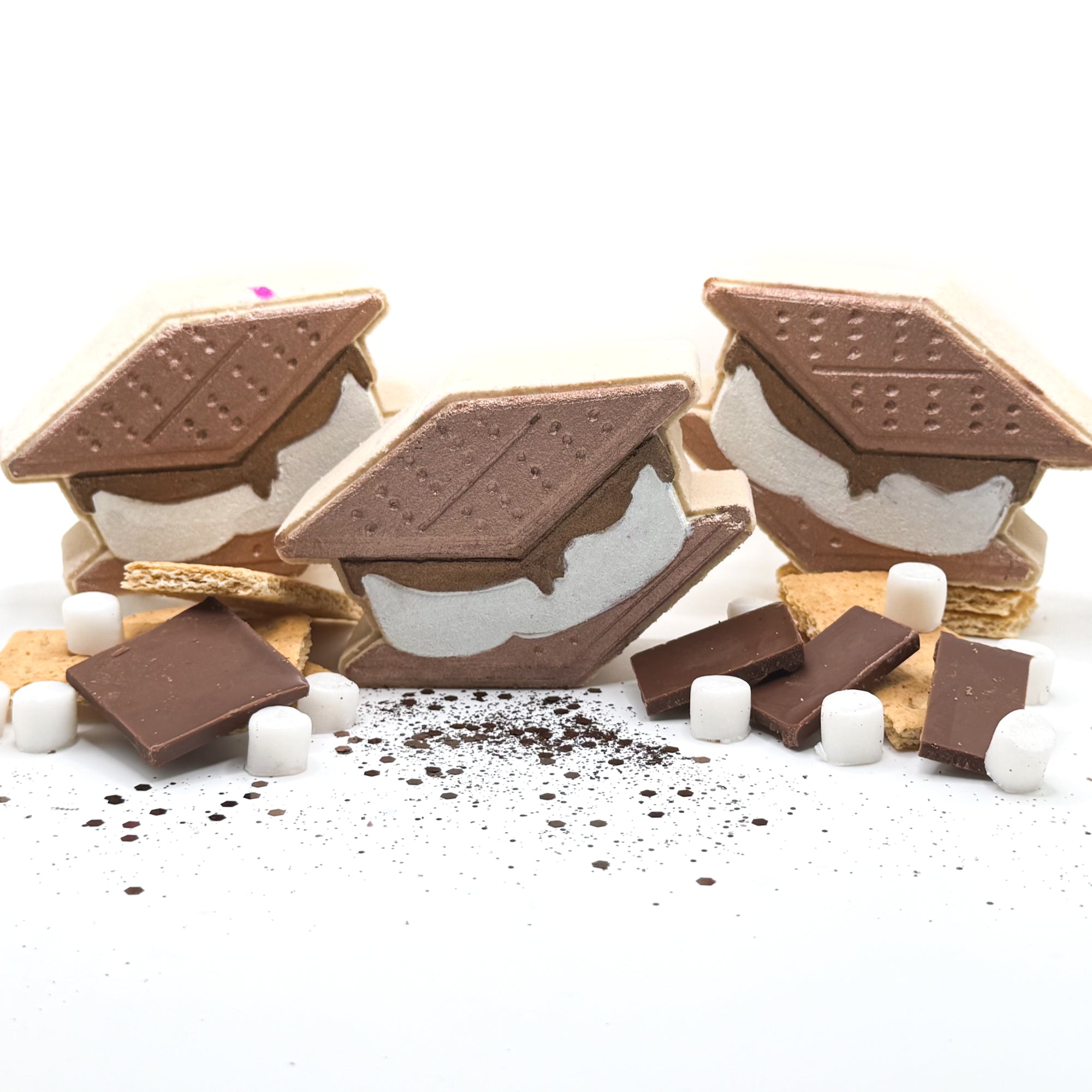 Three handmade s’mores shaped bath bombs painted to resemble a s’more with s’mores decorations around it on a white background.